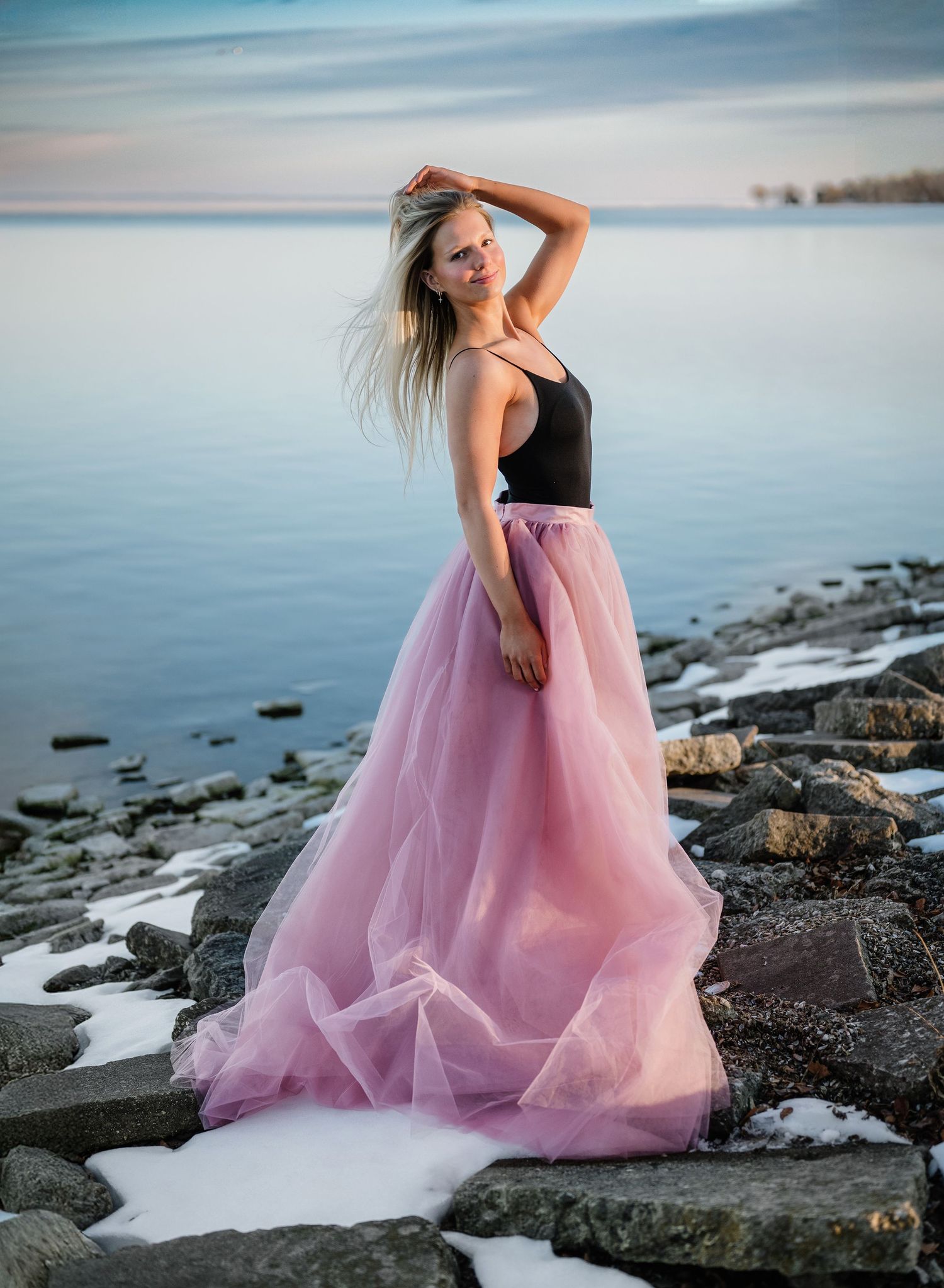 Blonde woman in black top and pink tulle skirt posing on rocky shoreline with snowy ground and calm water at sunset.