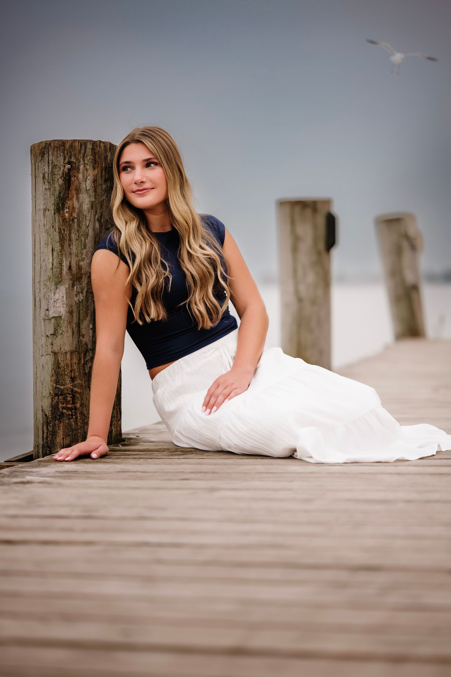 Young woman with long blonde hair in navy crop top and white skirt sitting on a wooden dock by the water.