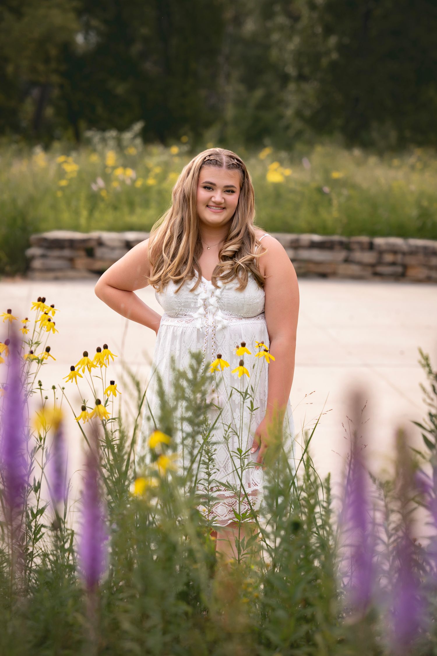 Young woman in white dress smiling among yellow and purple wildflowers in a garden setting.