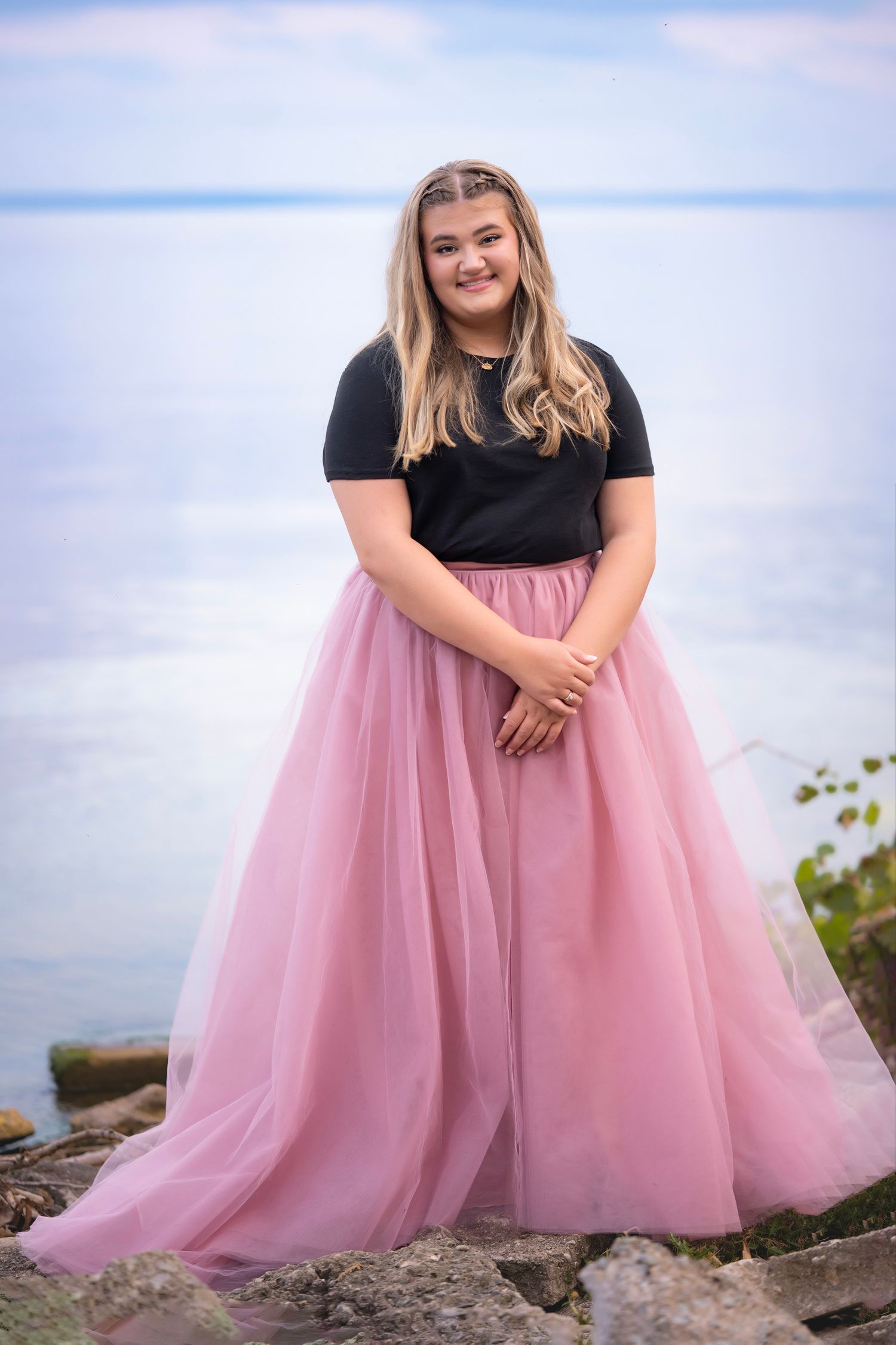 Young woman in black top and pink tulle skirt smiling by rocky lakeside.