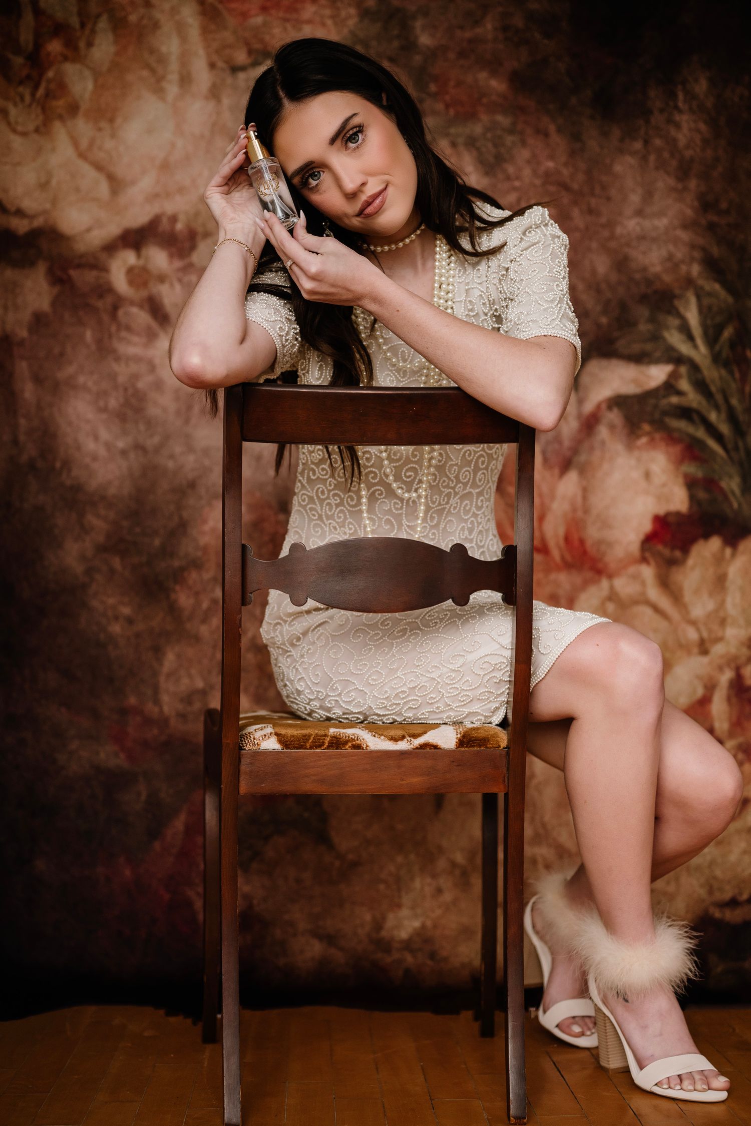 Young woman in white lace dress and pearl necklace poses elegantly on wooden chair against floral backdrop.