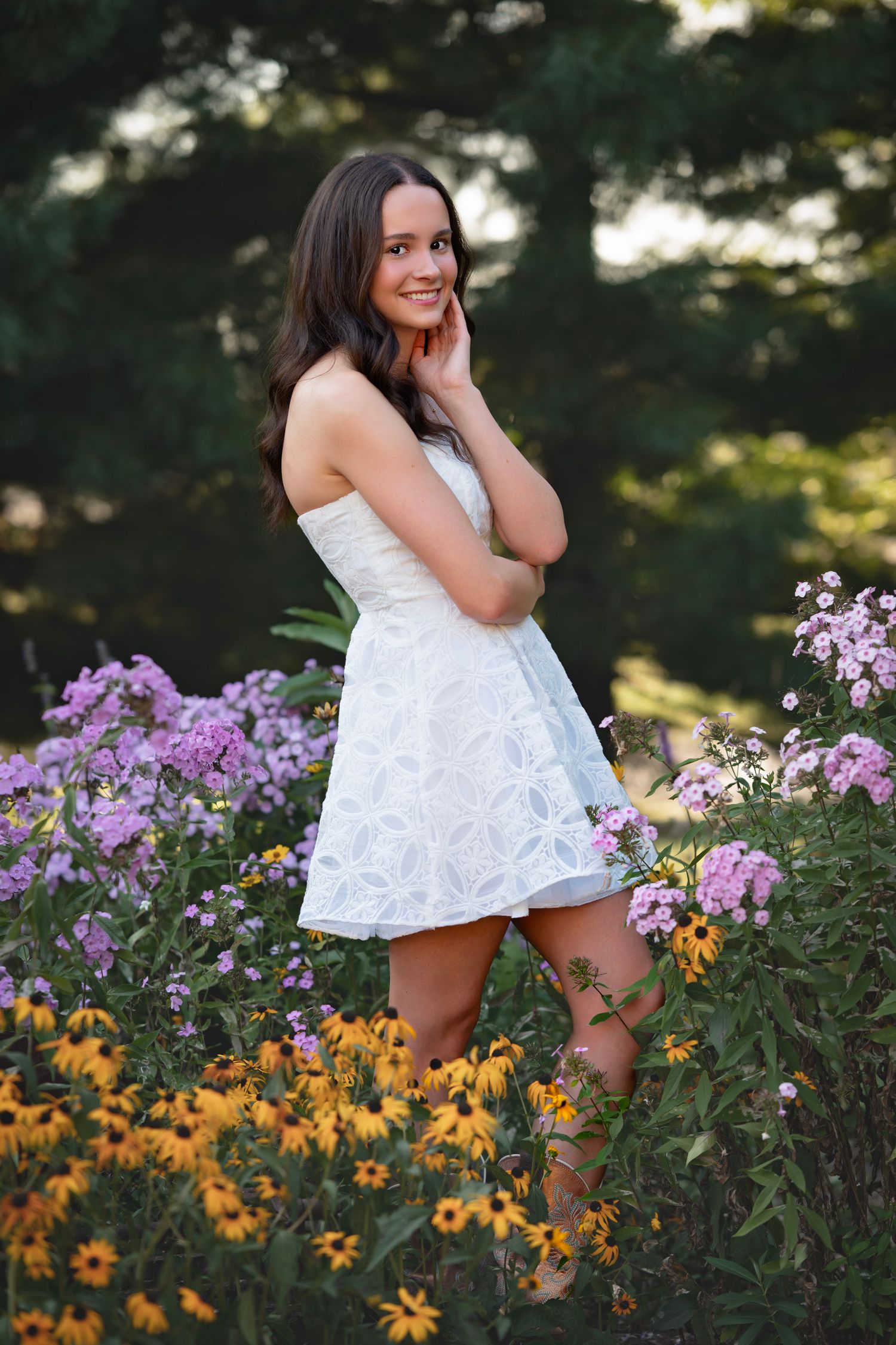 Young woman in white dress standing among colorful wildflowers, smiling outdoors in a lush garden setting.