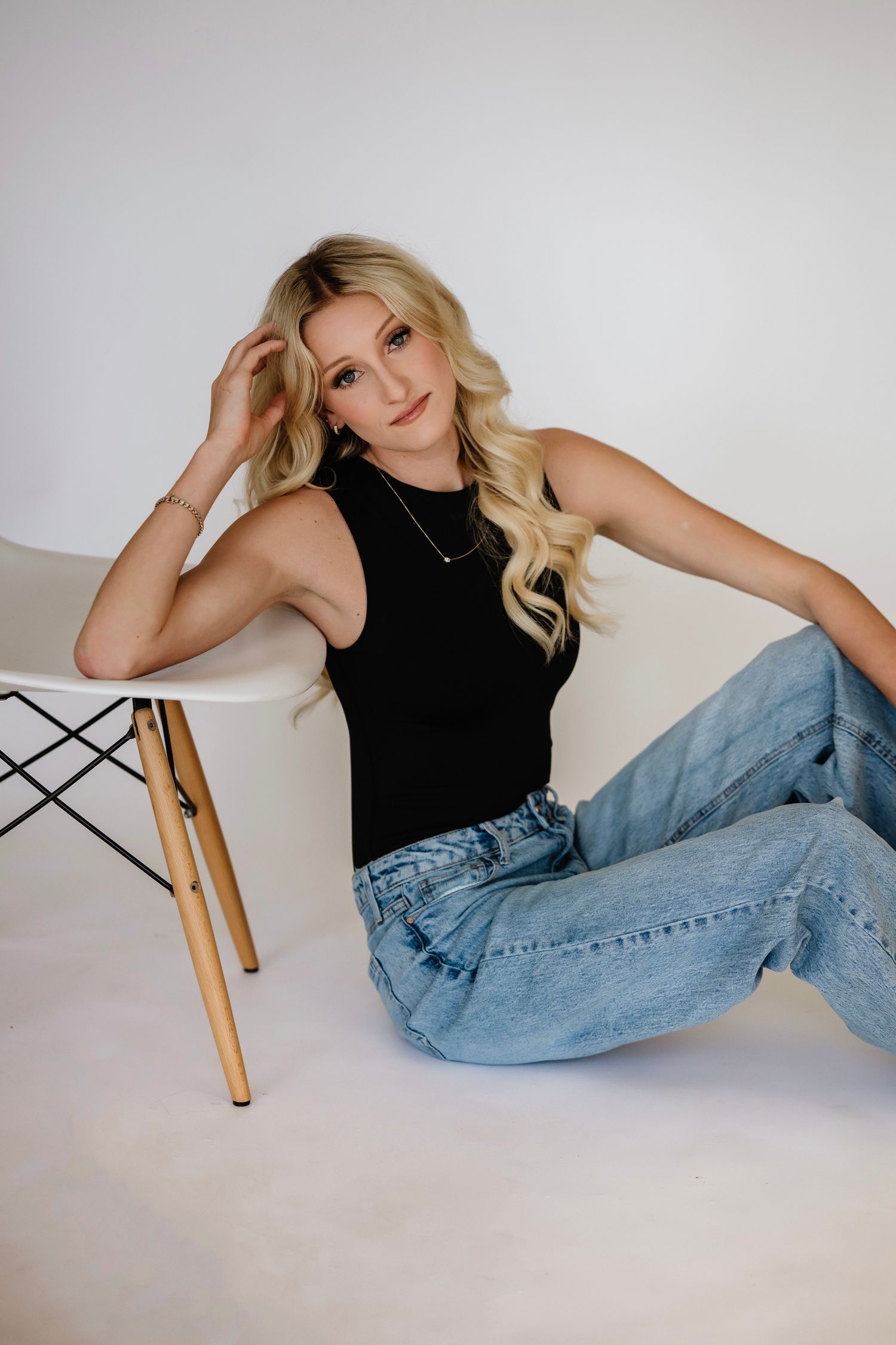 Blonde woman in black tank top and wide-leg jeans sits on floor leaning against modern white chair.
