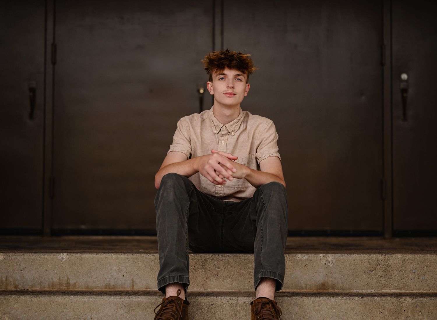 Young man with curly hair sitting on steps, wearing a beige polo shirt and jeans, posing confidently outdoors.