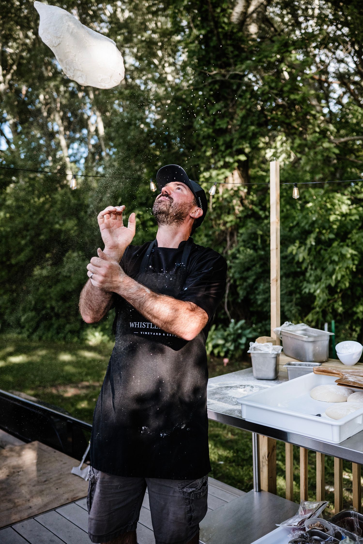 Chef tossing pizza dough in the air outdoors, wearing a black apron dusted with flour near a prep table.