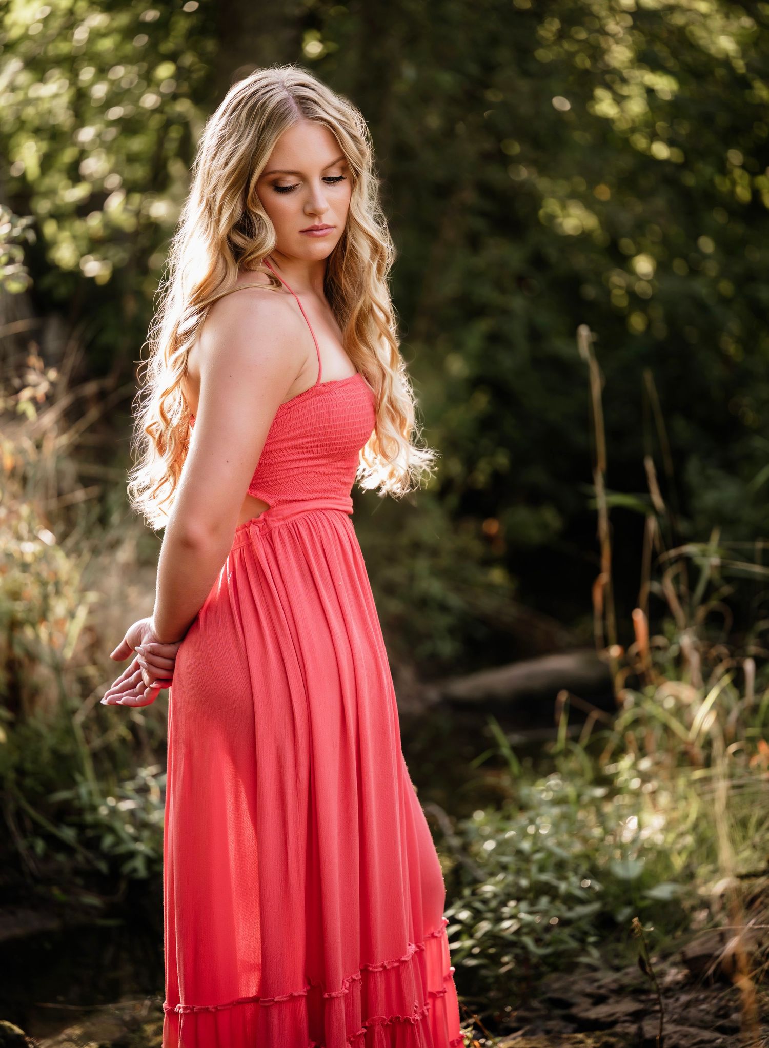 A woman in a flowing coral maxi dress poses gracefully in a sunlit forest setting with glowing golden light.