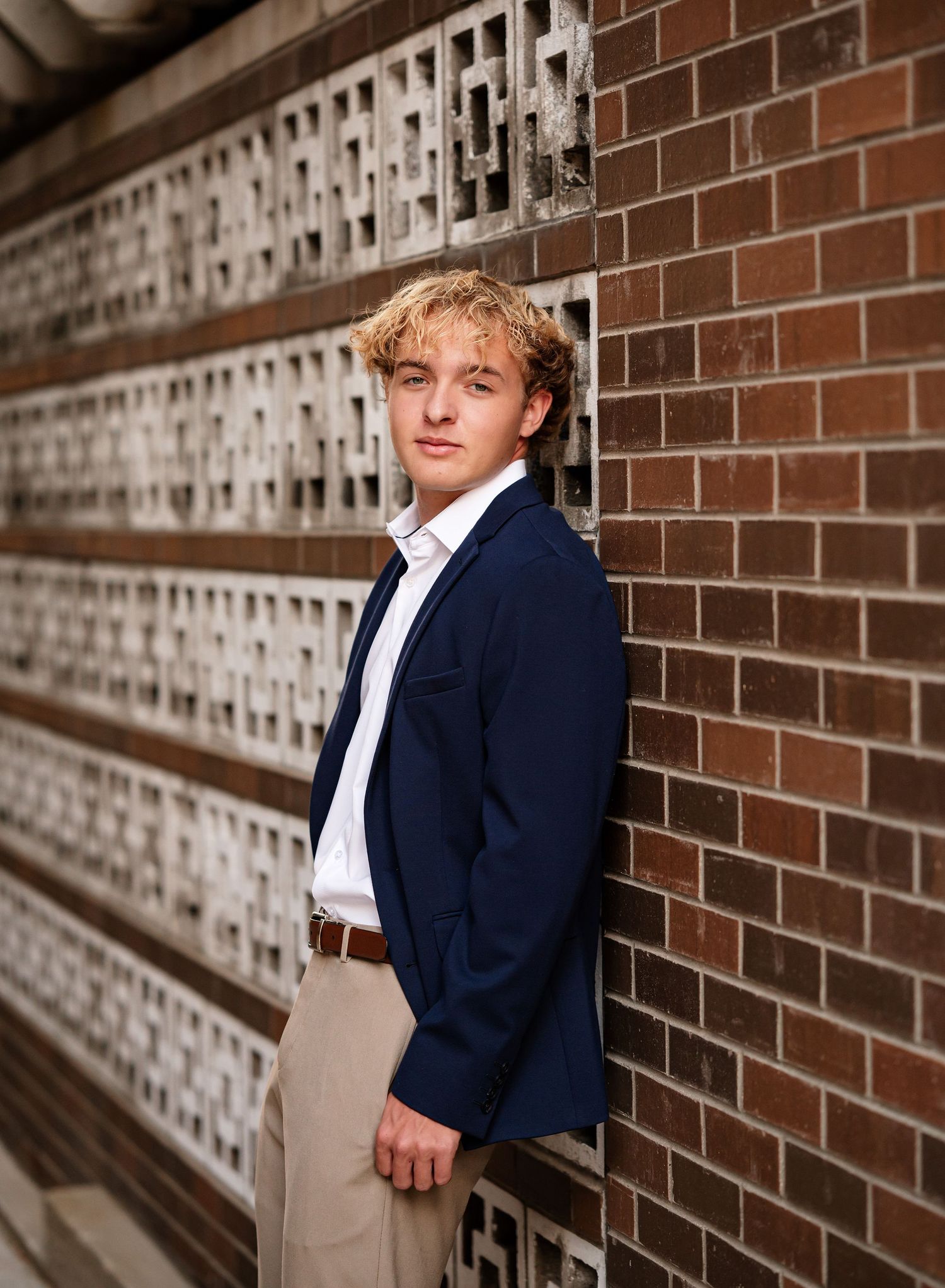 Someone in a navy blazer and khakis poses against a decorative brick wall.