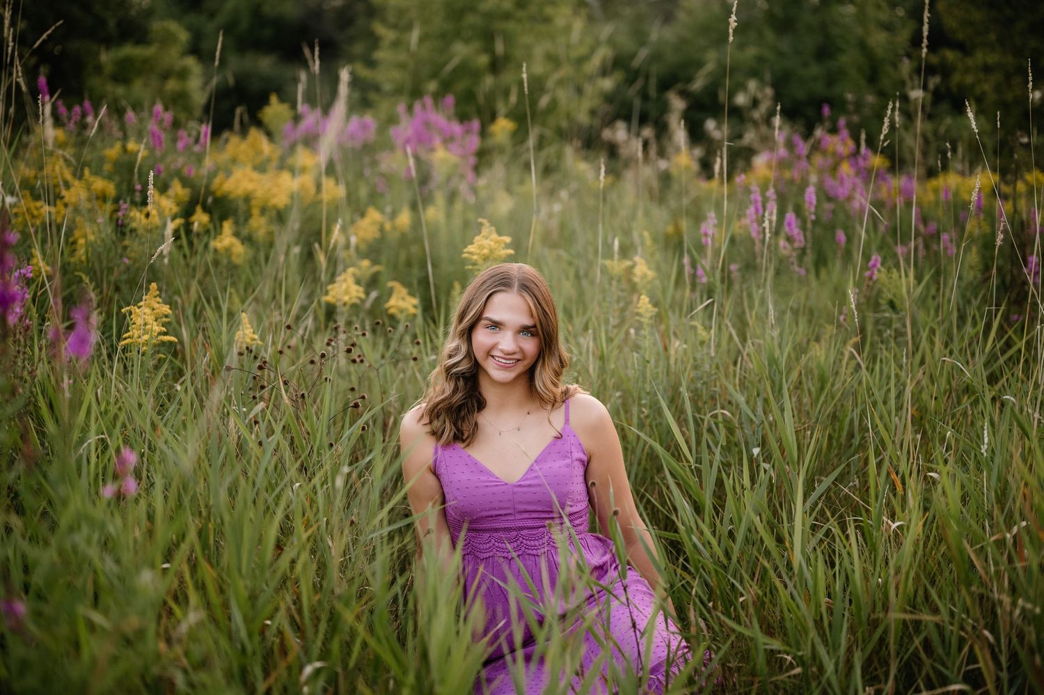 Senior portrait in a purple dress among tall wildflowers and natural grasses.