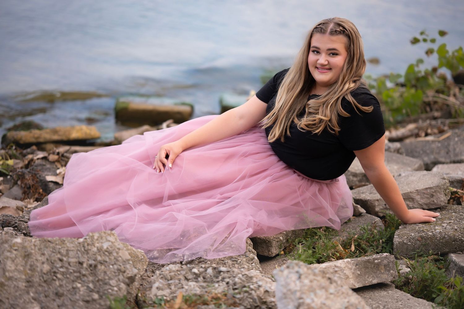 Young girl in pink tulle skirt and black top poses on rocky shoreline with water in background.