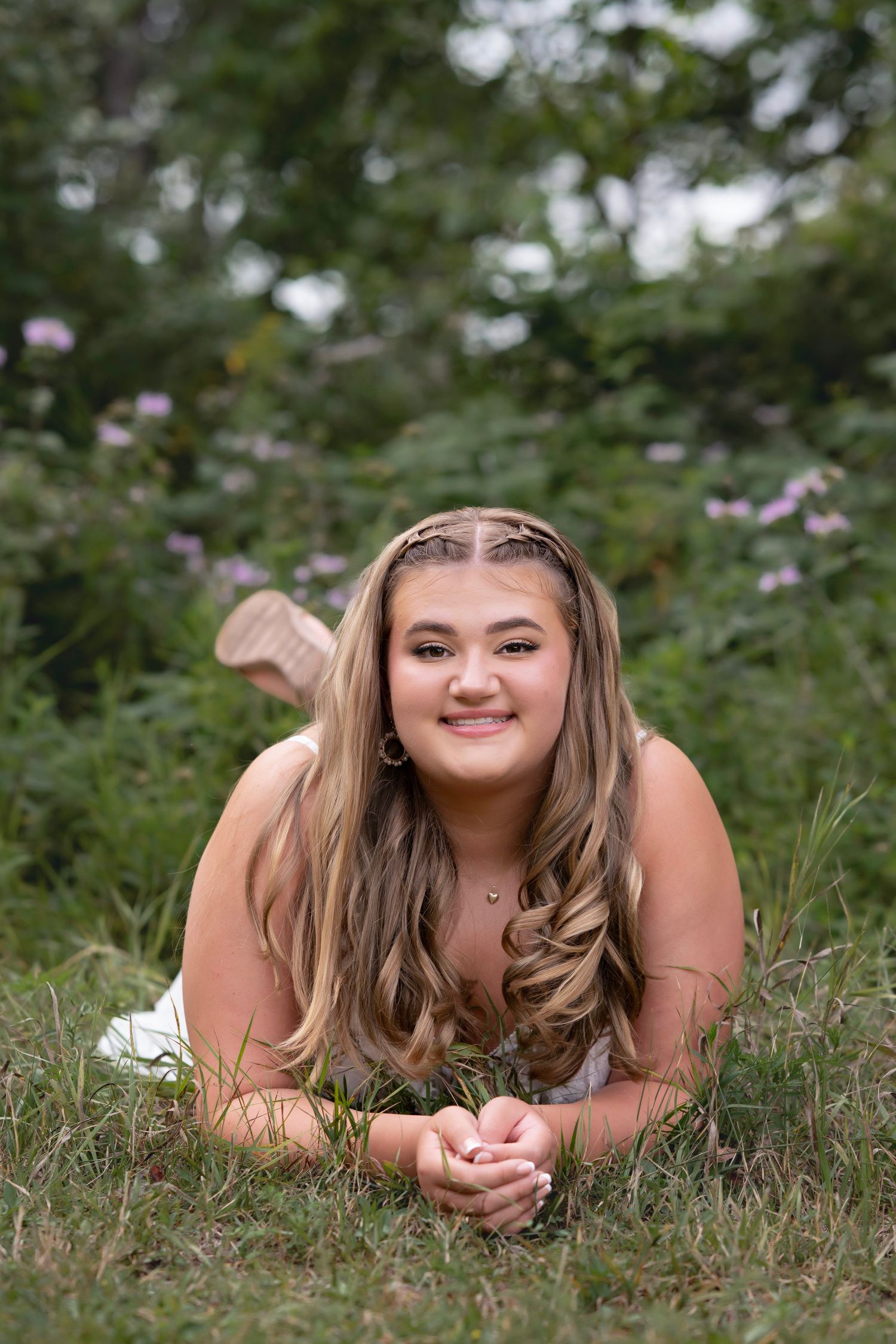Young woman with long wavy hair lying on grass, smiling, surrounded by greenery and pink flowers outdoors.