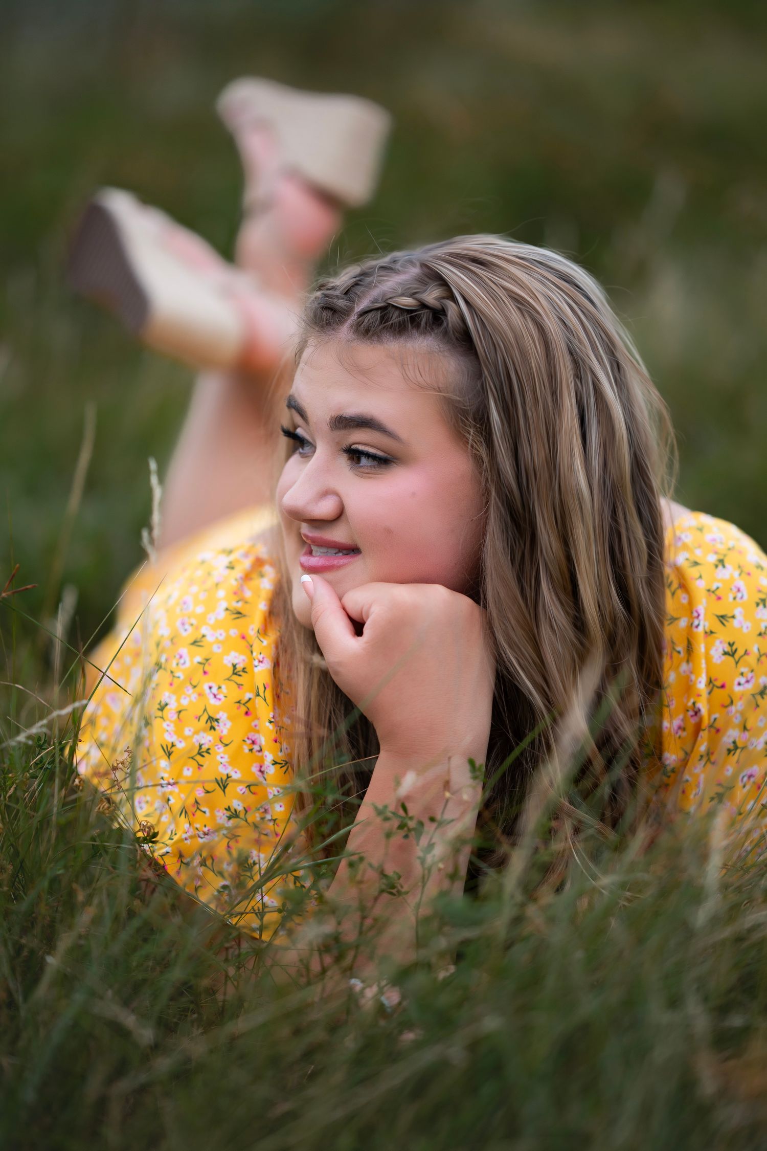 Teen girl in yellow floral dress lying in grass, resting chin on hand, smiling softly outdoors.