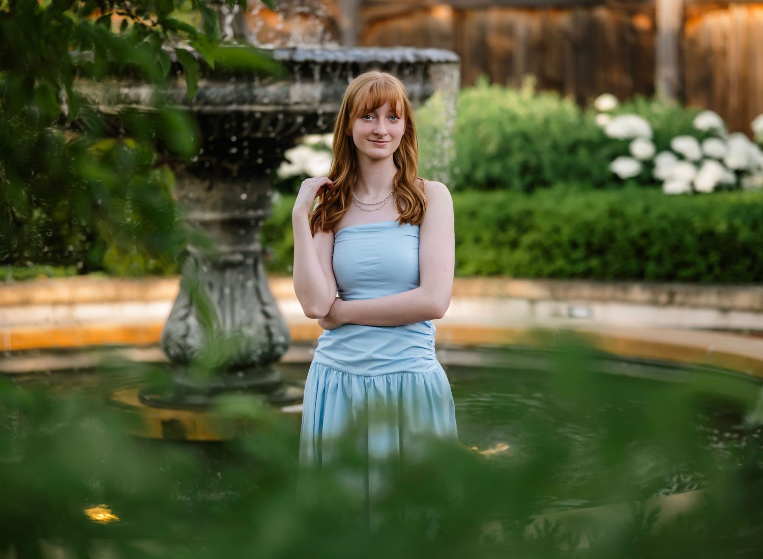 A person in a light blue dress stands by a tranquil garden pond surrounded by lush greenery on a sunny day.
