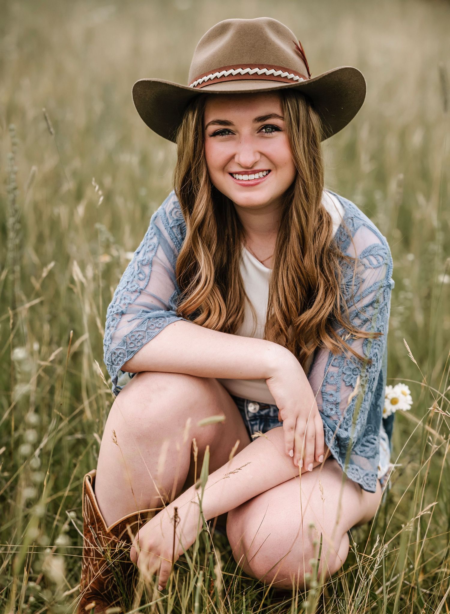 A western-style portrait in a field wearing a brown cowboy hat, denim jacket and white top while sitting in tall grass.