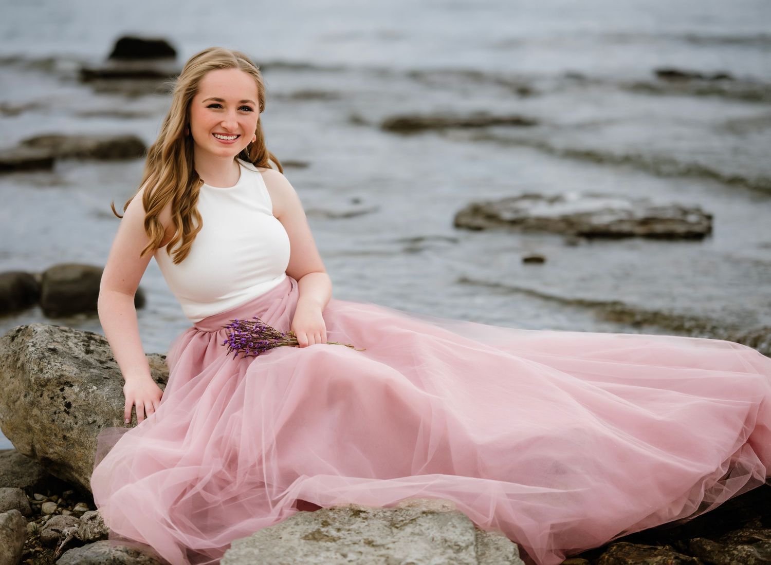 A flowing pink tulle dress billows in the wind against a rocky beach backdrop during a seaside photoshoot.