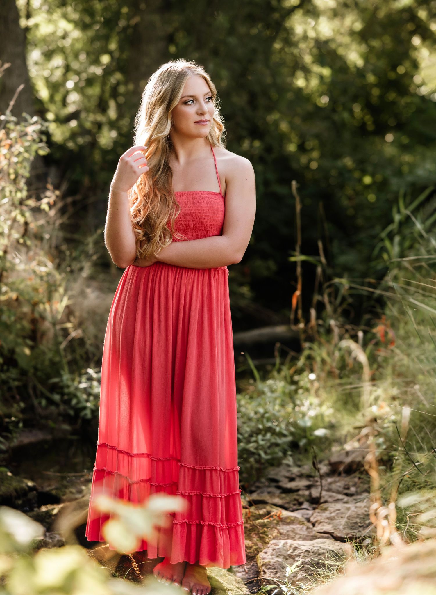 Young woman in coral maxi dress poses gracefully among lush green trees and foliage in an outdoor portrait.