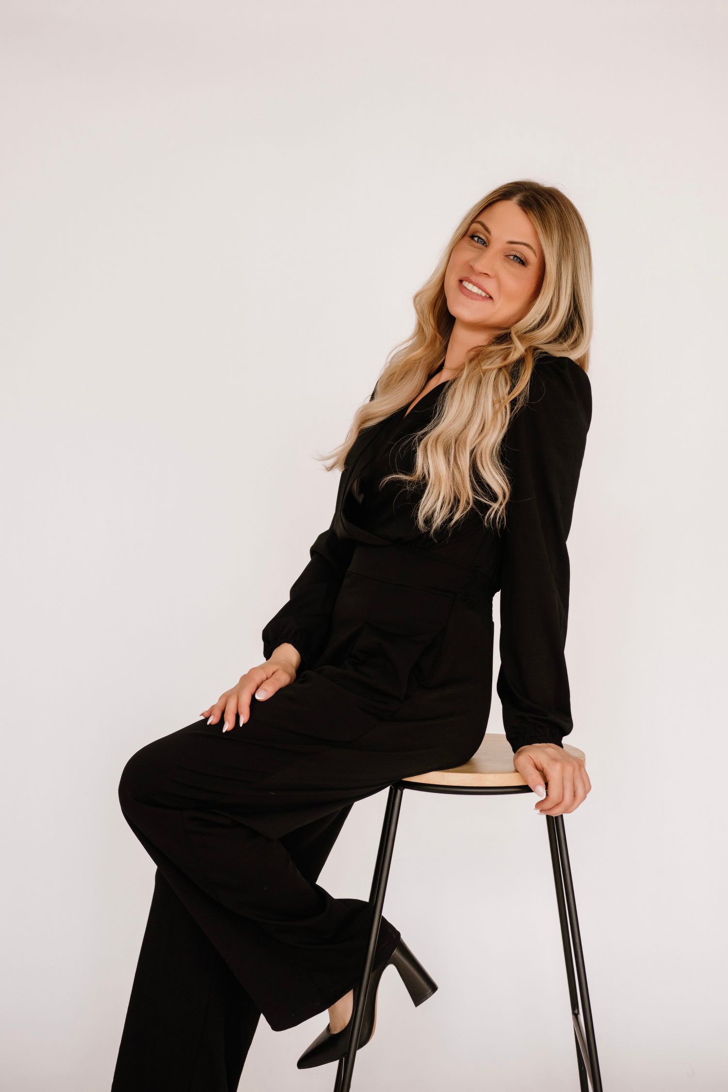 Stylish black long sleeve jumpsuit with wide legs and fitted waist worn with heels while sitting on stool against white backdrop.