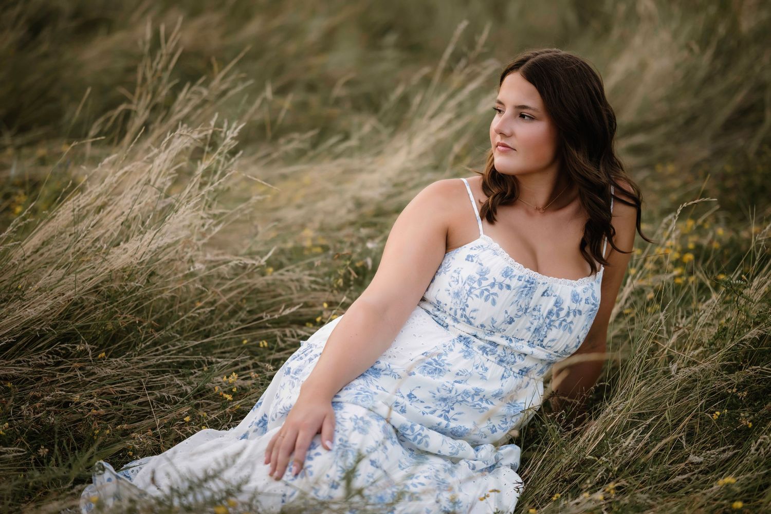 A person in a floral blue and white sundress sits among tall grass in a dreamy outdoor setting.