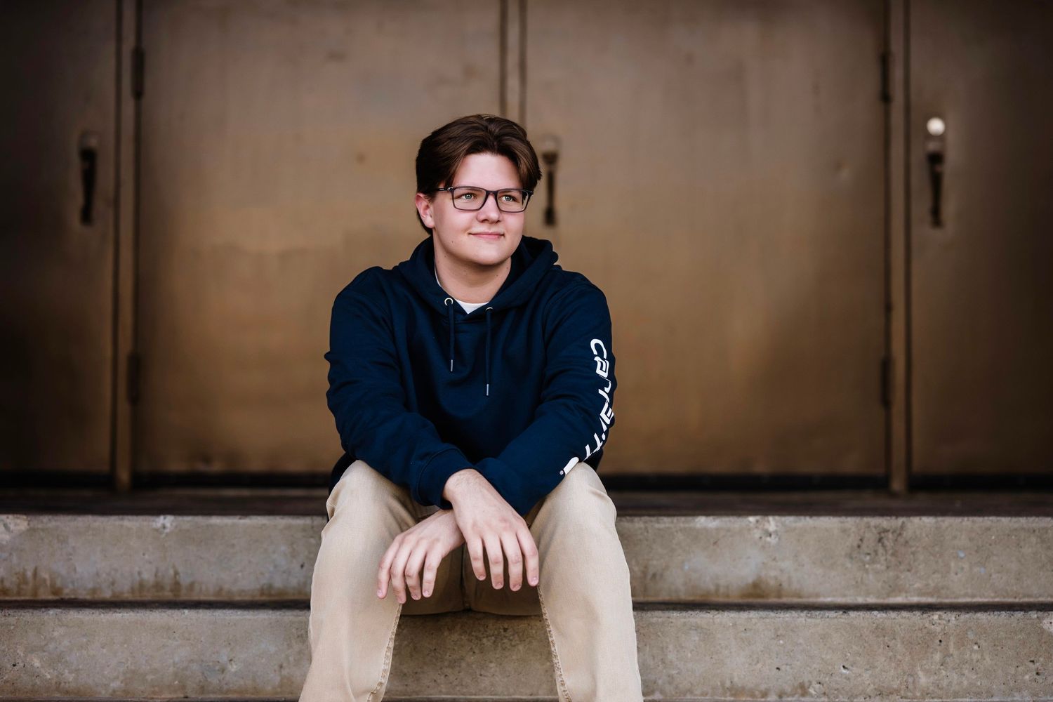 A person in a navy blue hoodie and khaki pants sits on a concrete step in an industrial hallway setting.