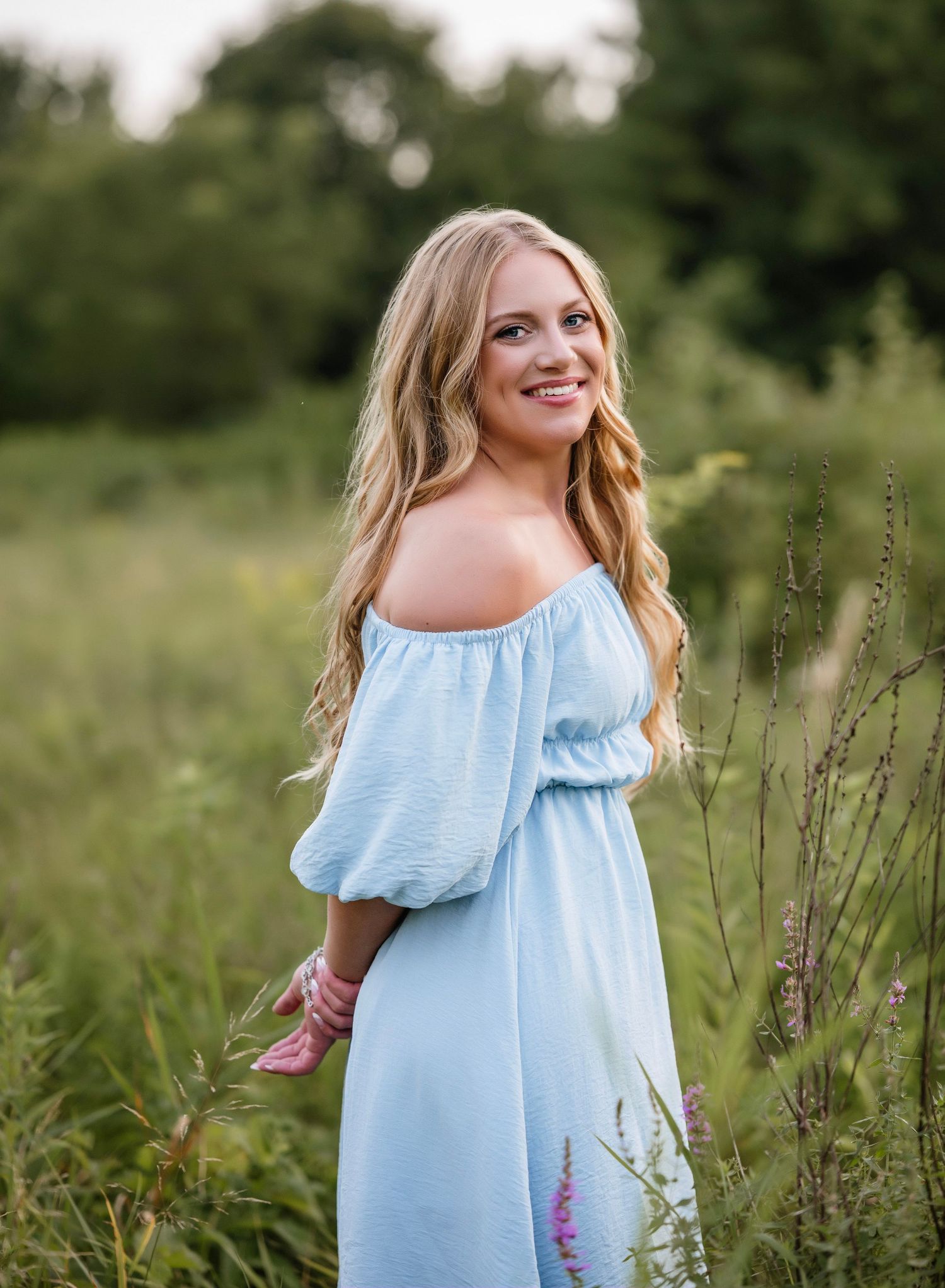 A woman in a light blue off-shoulder dress poses gracefully in a field of tall grass and wildflowers.