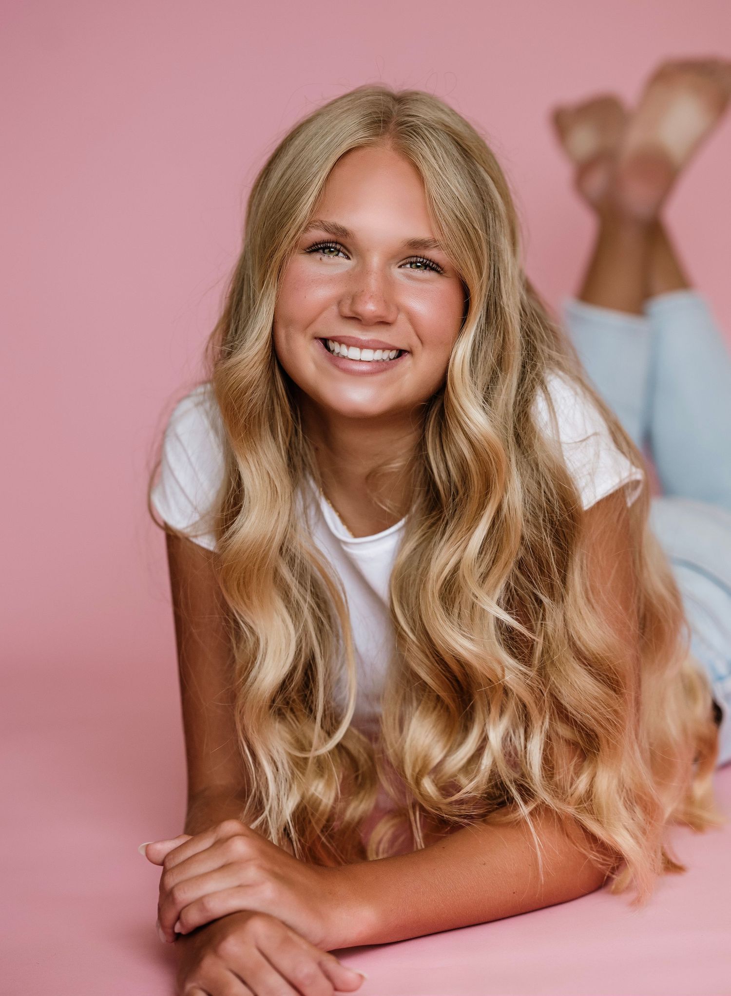 Woman with wavy blonde hair poses against pink background in casual white top.