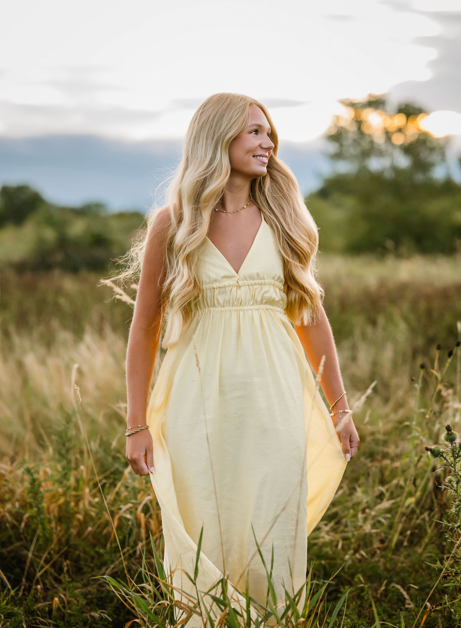A person in a flowing yellow sundress stands in a sunlit meadow of tall grass during golden hour.