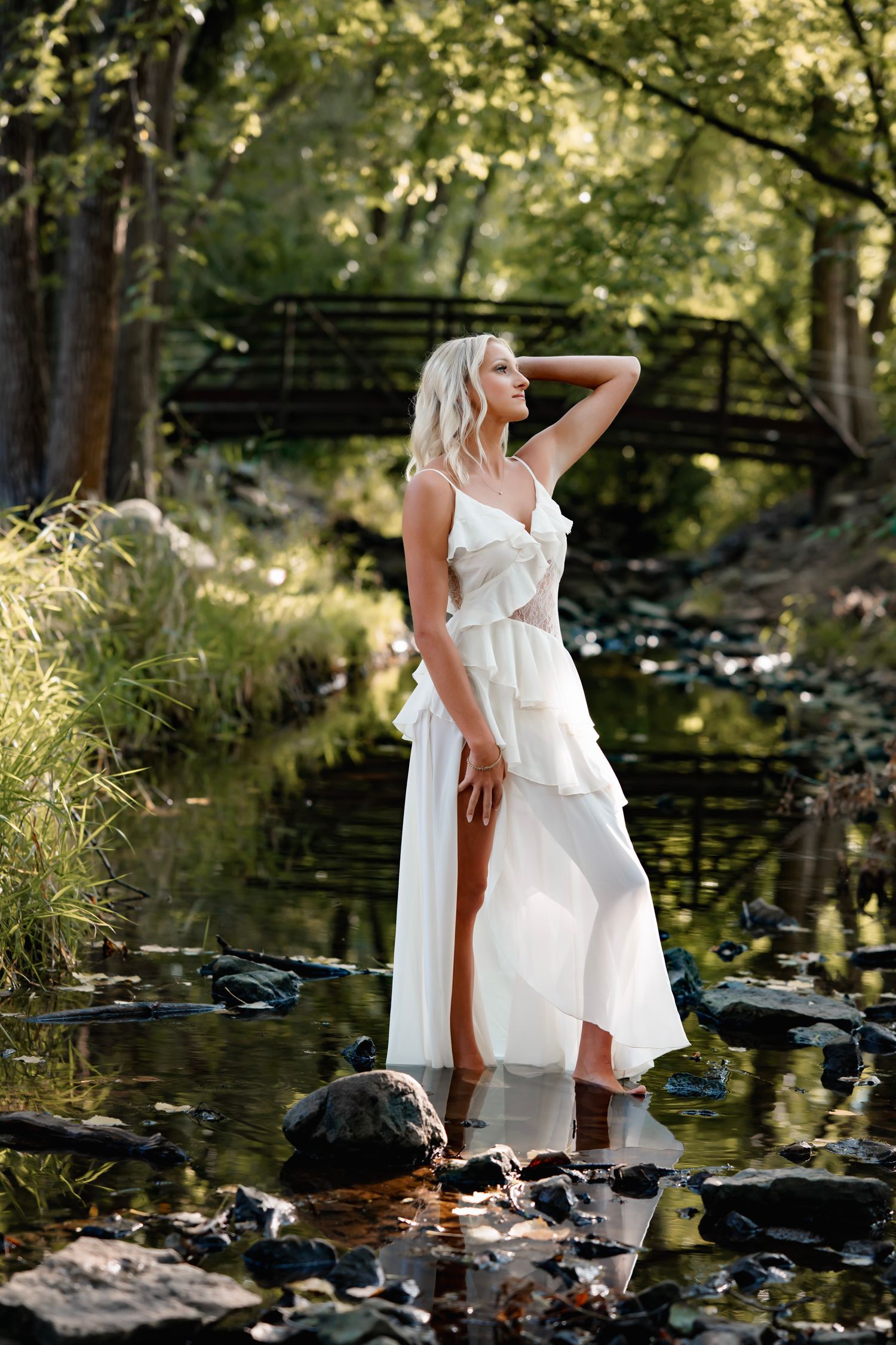Woman in flowing white dress poses gracefully by a tranquil pond surrounded by rocks and trees in natural light.