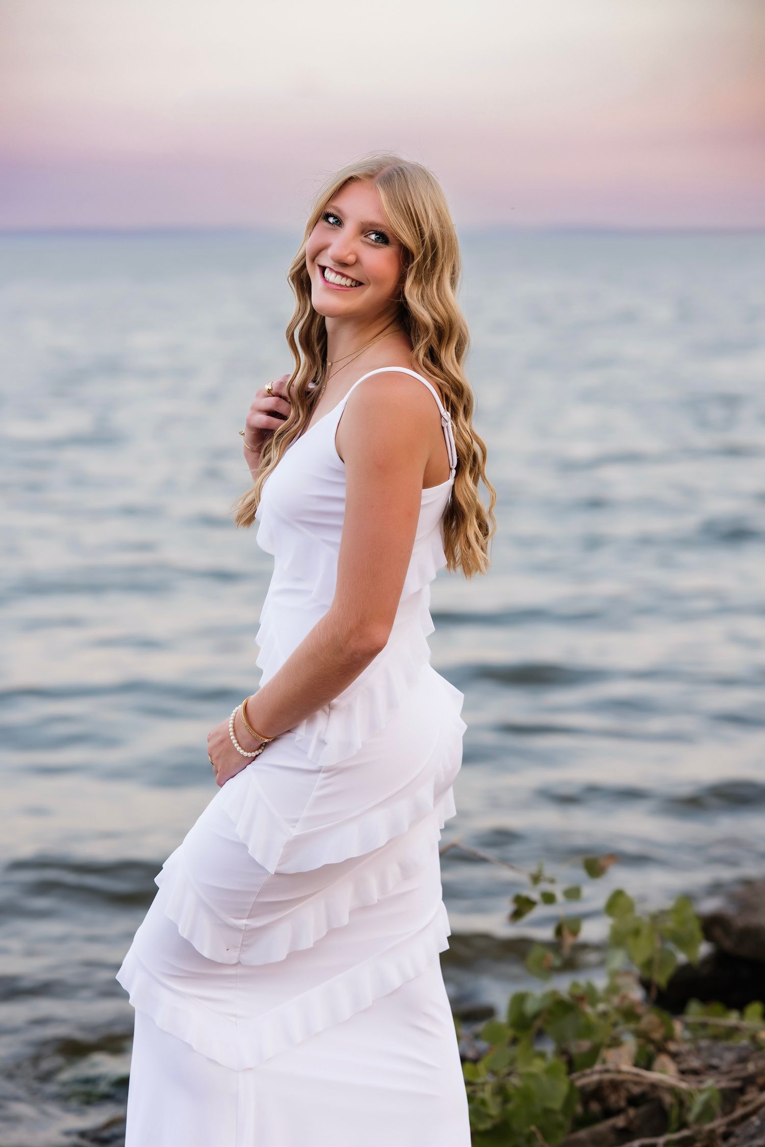 Young blonde woman in white ruffled dress smiling at beach during sunset.