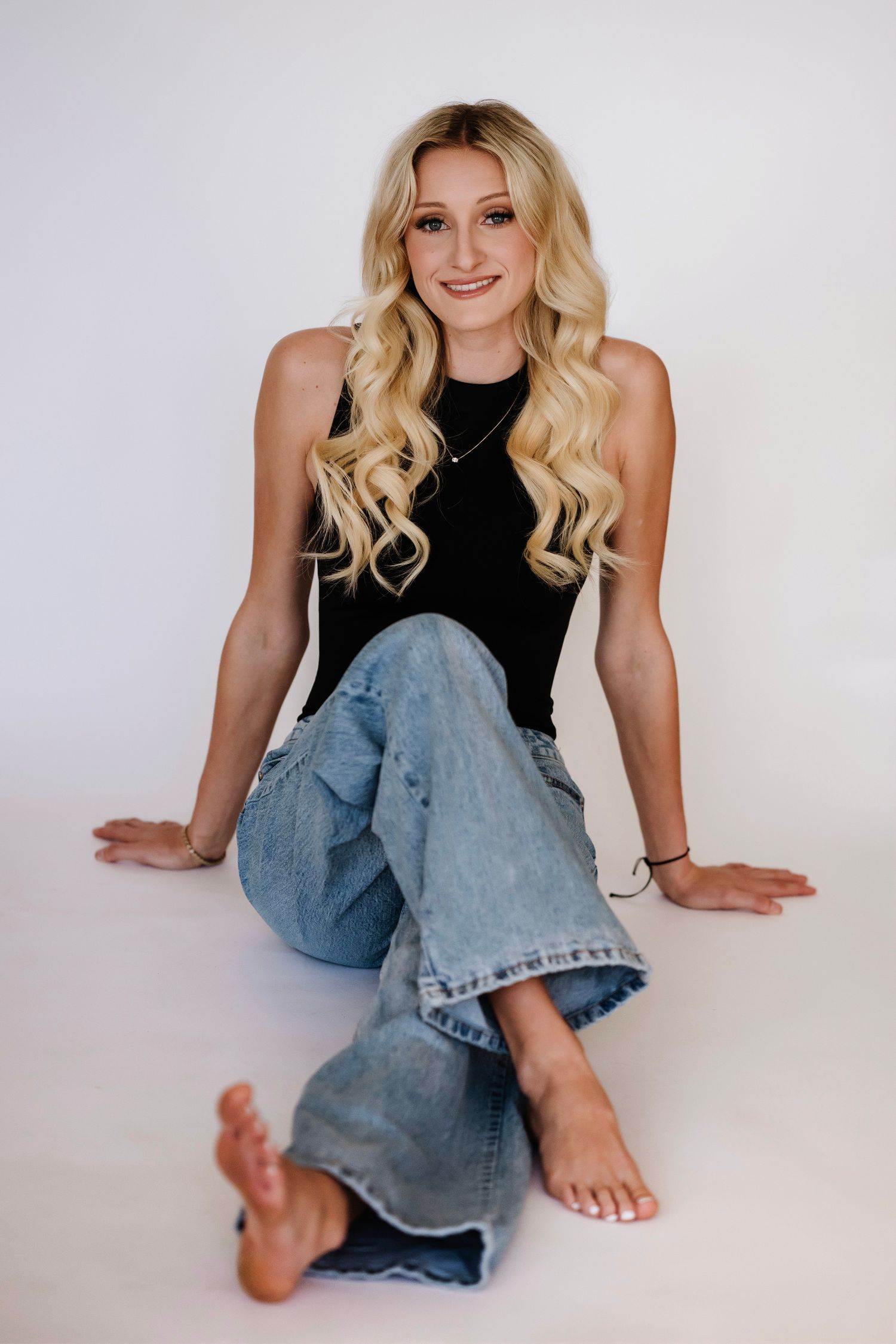 Smiling blonde woman sitting on white floor in black tank top and wide-leg jeans, barefoot, with wavy hair.