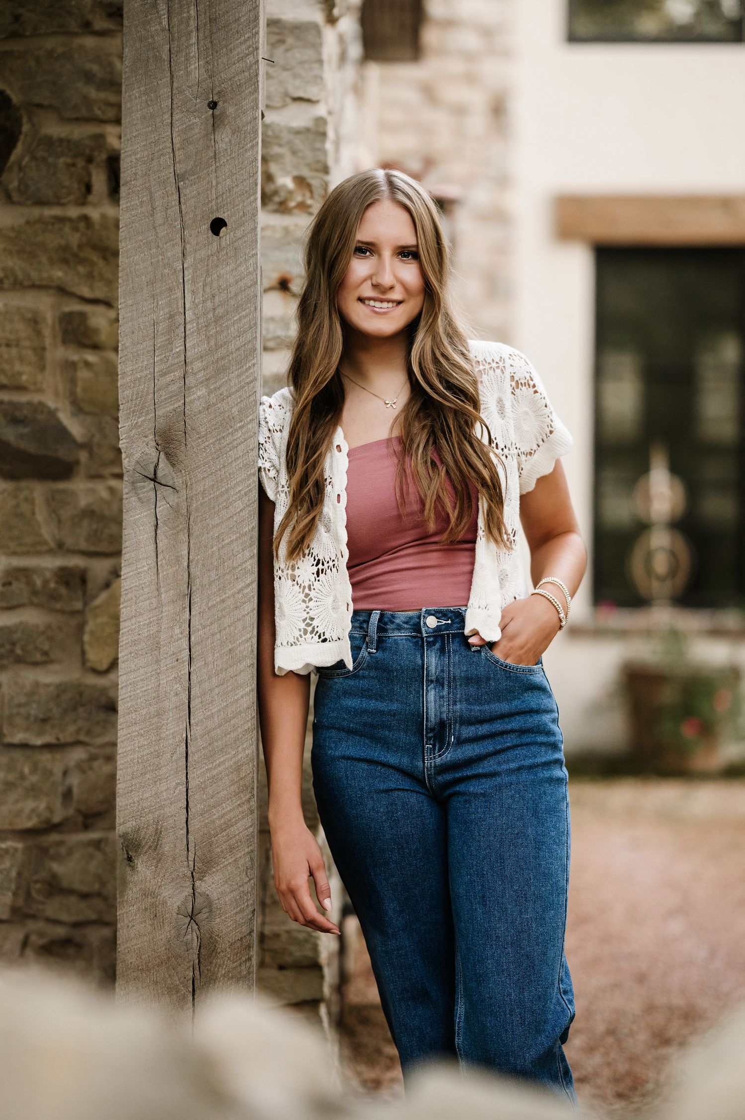 Young woman in crochet vest, pink top, and wide-leg jeans poses by rustic wood post and stone wall.