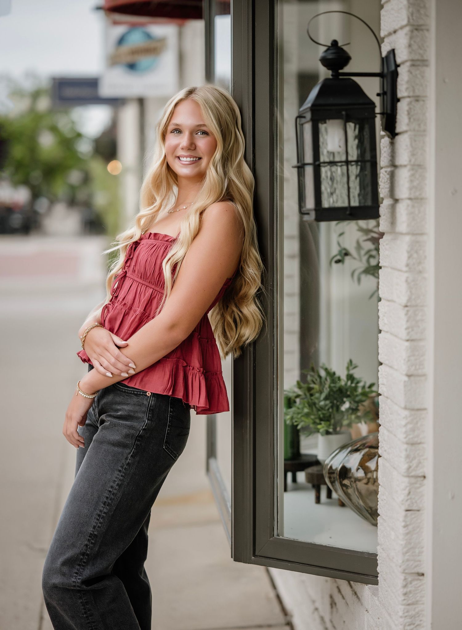 Young blonde woman in red top and black jeans leaning against a white brick storefront with a lantern.