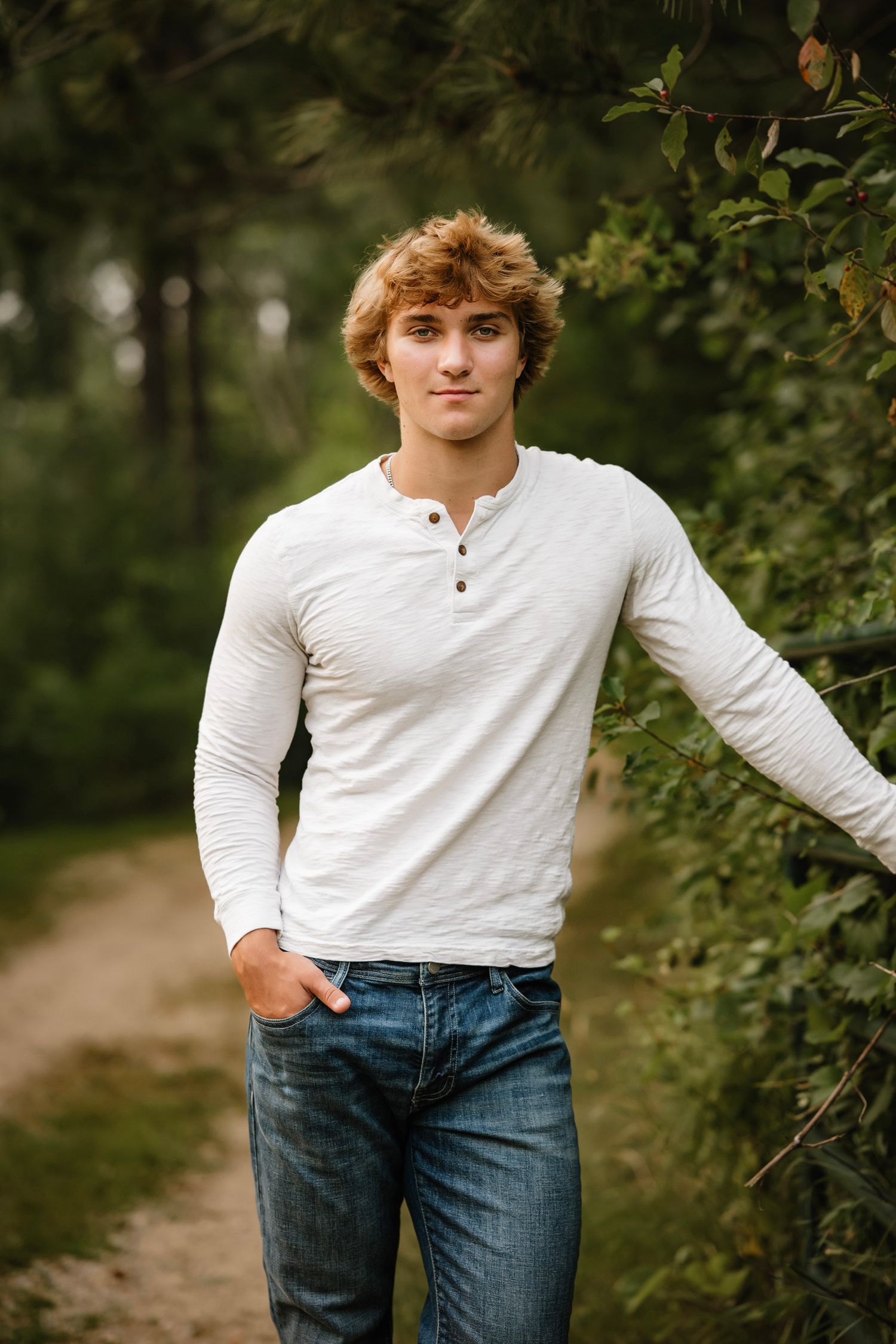 A person in a white long-sleeve henley shirt and blue jeans poses casually against a natural outdoor background.