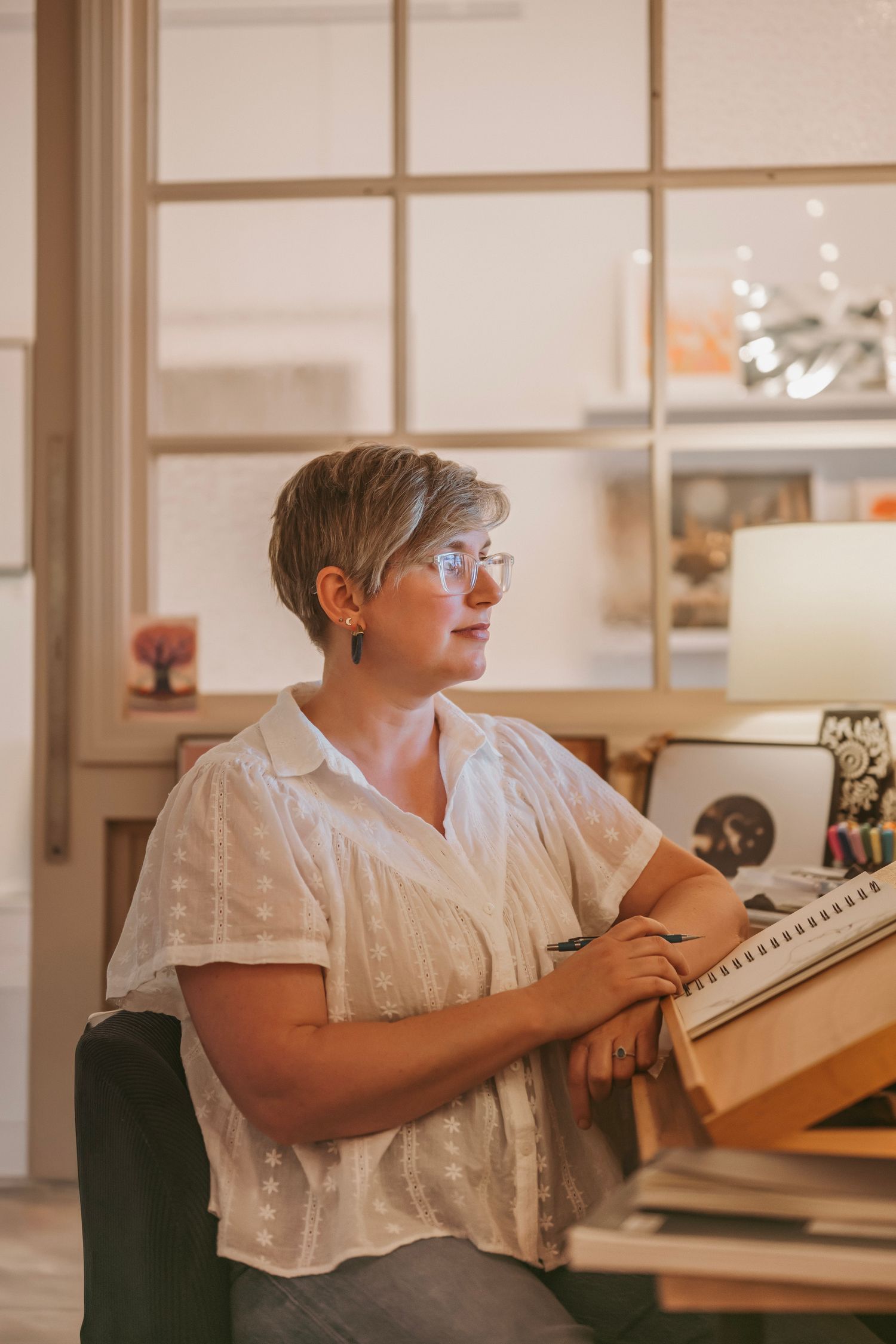 A person in a white blouse sits at a desk with books, wearing glasses and looking thoughtfully to the side.