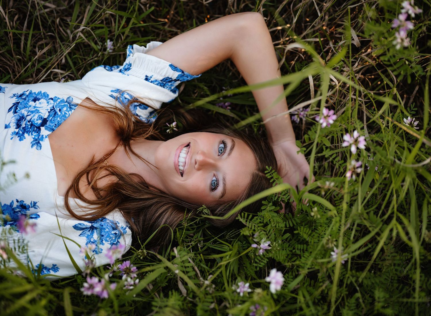 Artistic senior portrait lying in grass with blue wildflowers creating a dreamy scene.