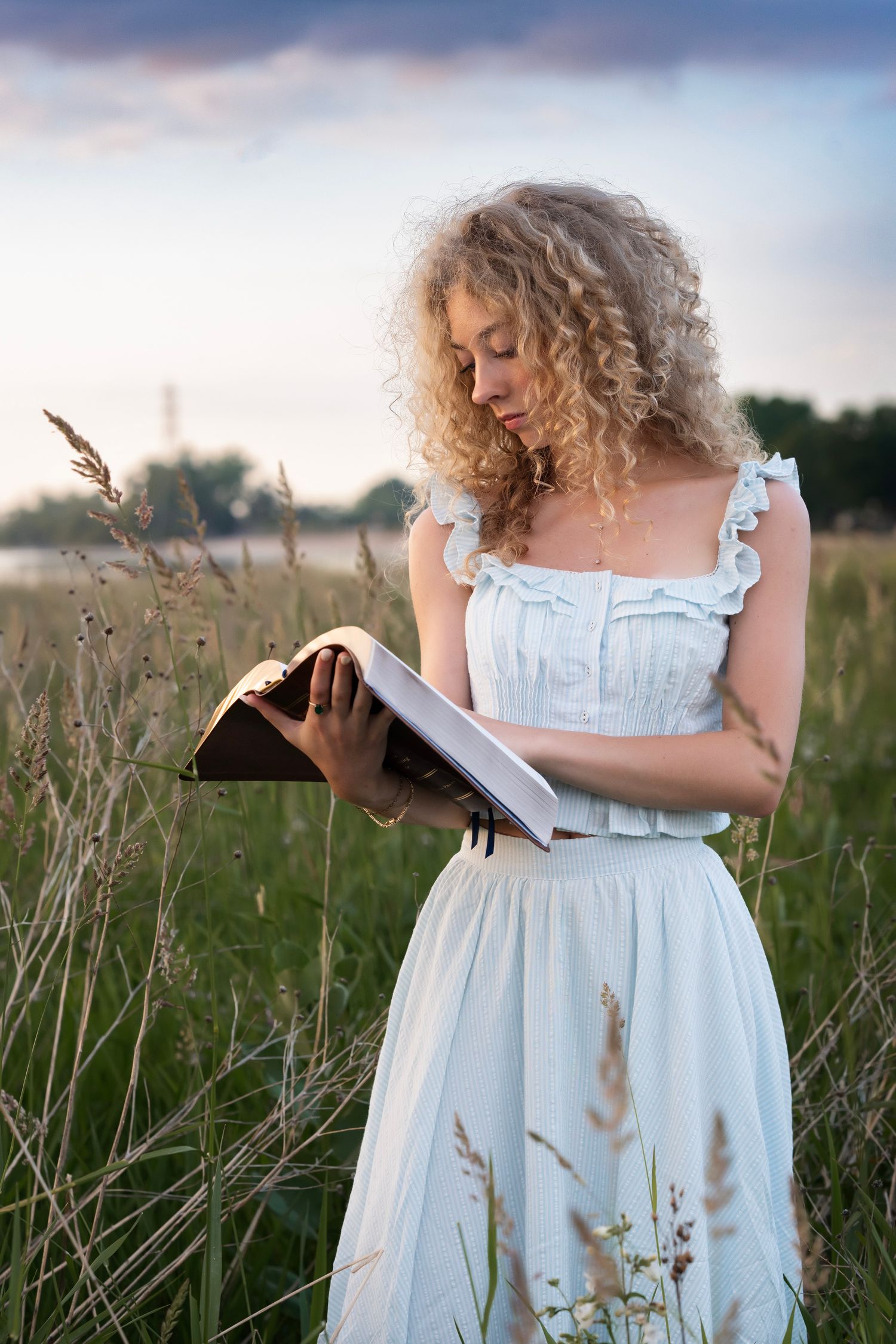Young woman with curly blonde hair reads a book in a field wearing a white dress at sunset.
