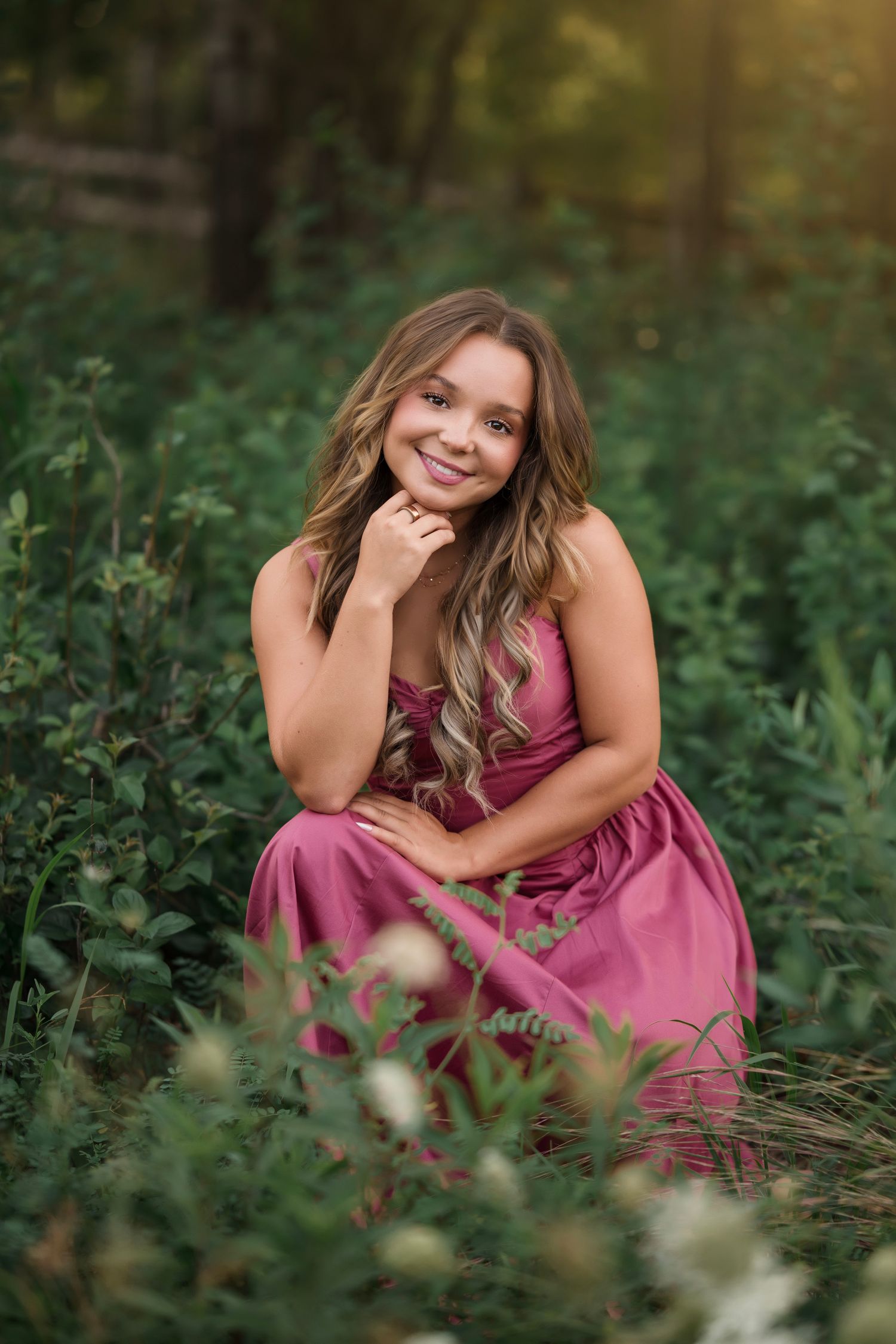 Young woman in pink dress sitting among green foliage, smiling outdoors in natural light.