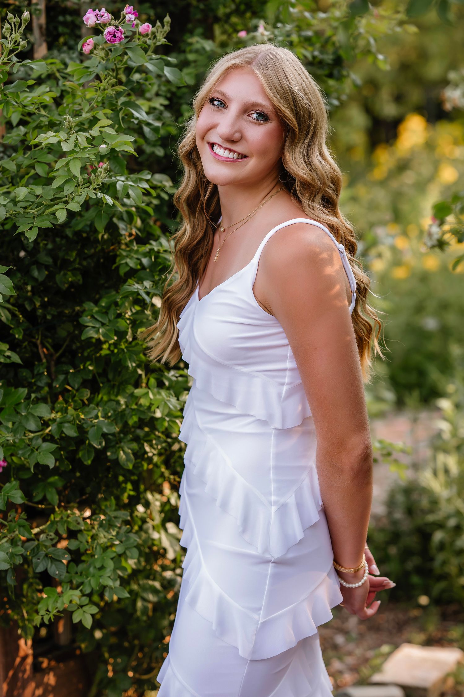 Young woman in white ruffled dress smiling in a lush garden with pink roses and greenery.