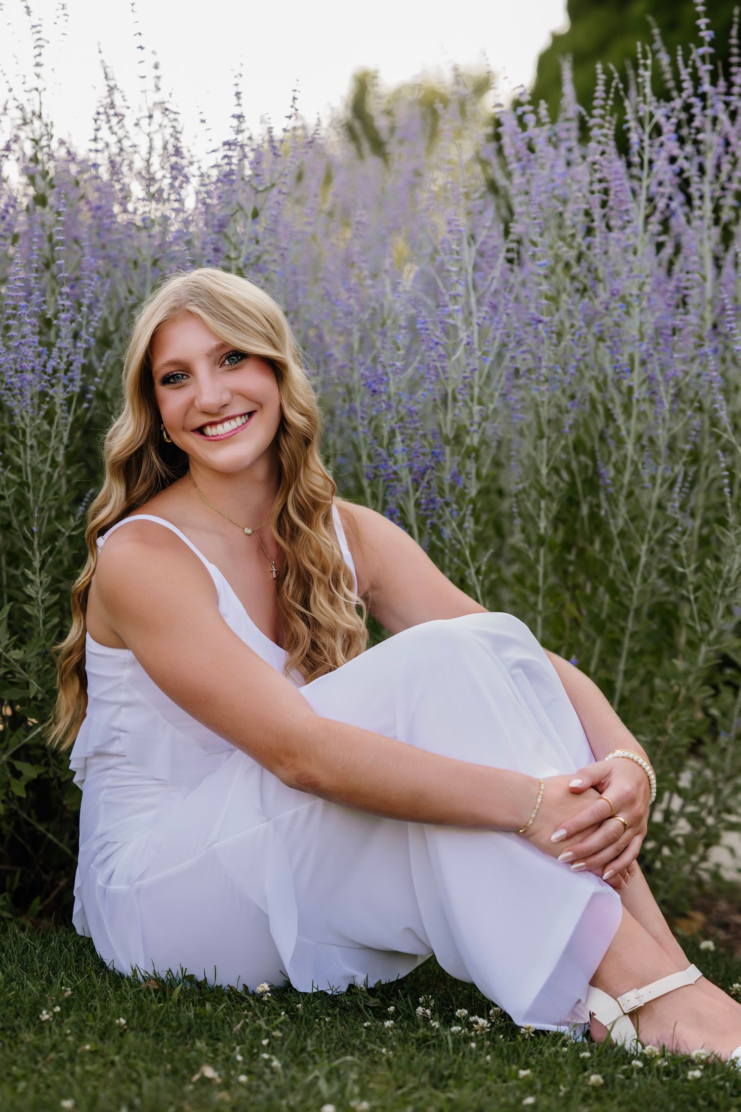 Young woman in white outfit sitting in front of lavender flowers, smiling outdoors.