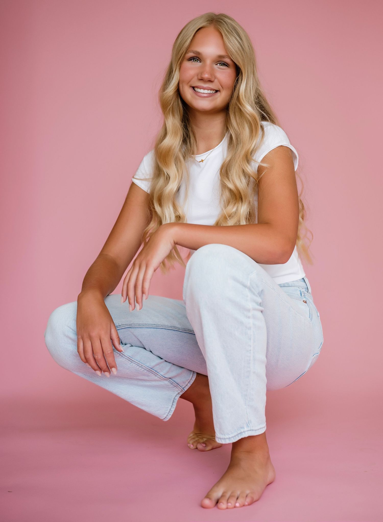 Smiling blonde woman in white tee and light jeans crouching barefoot against a pink background.