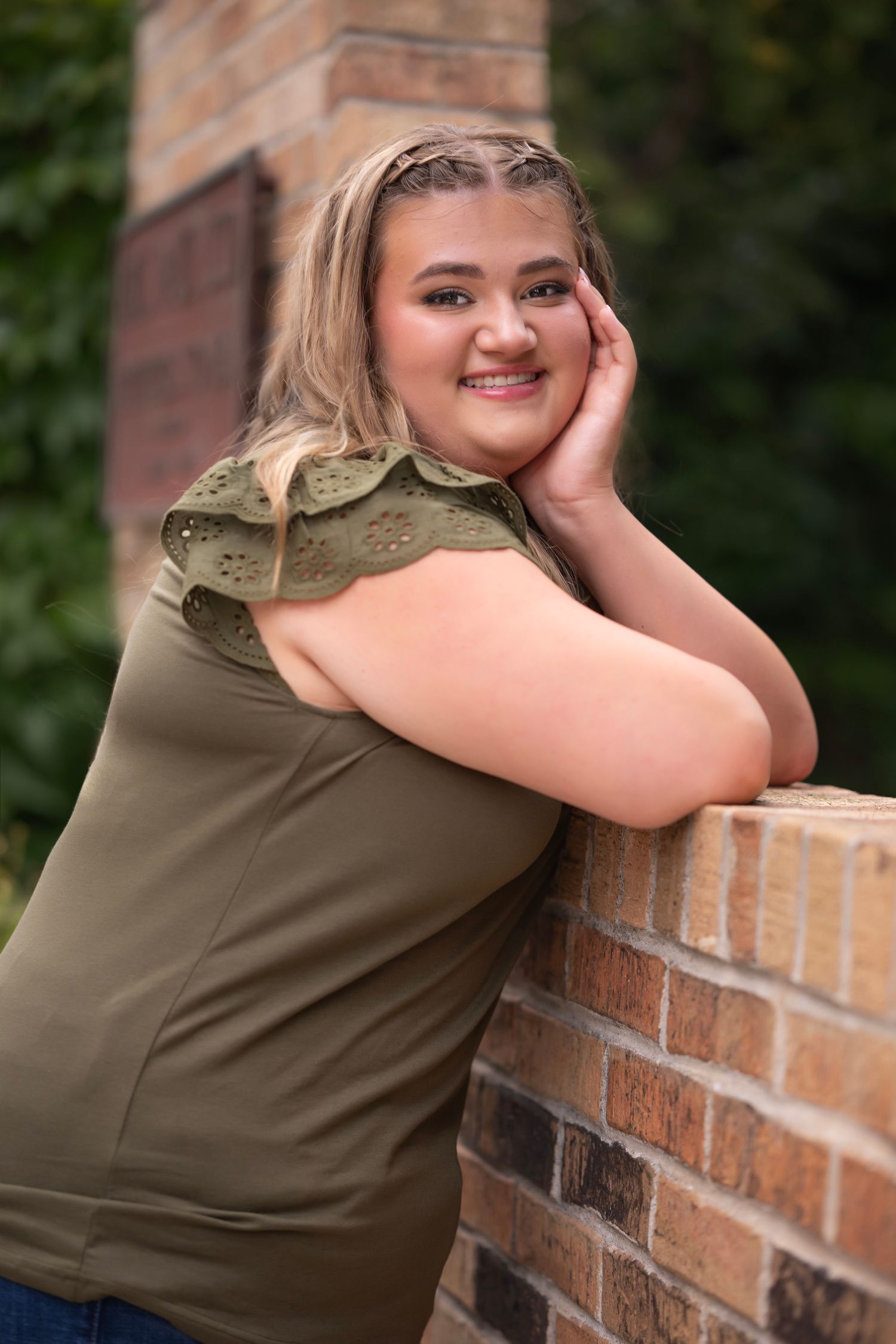 Smiling teenage girl in olive green top leaning on brick wall outdoors for senior portrait photo.