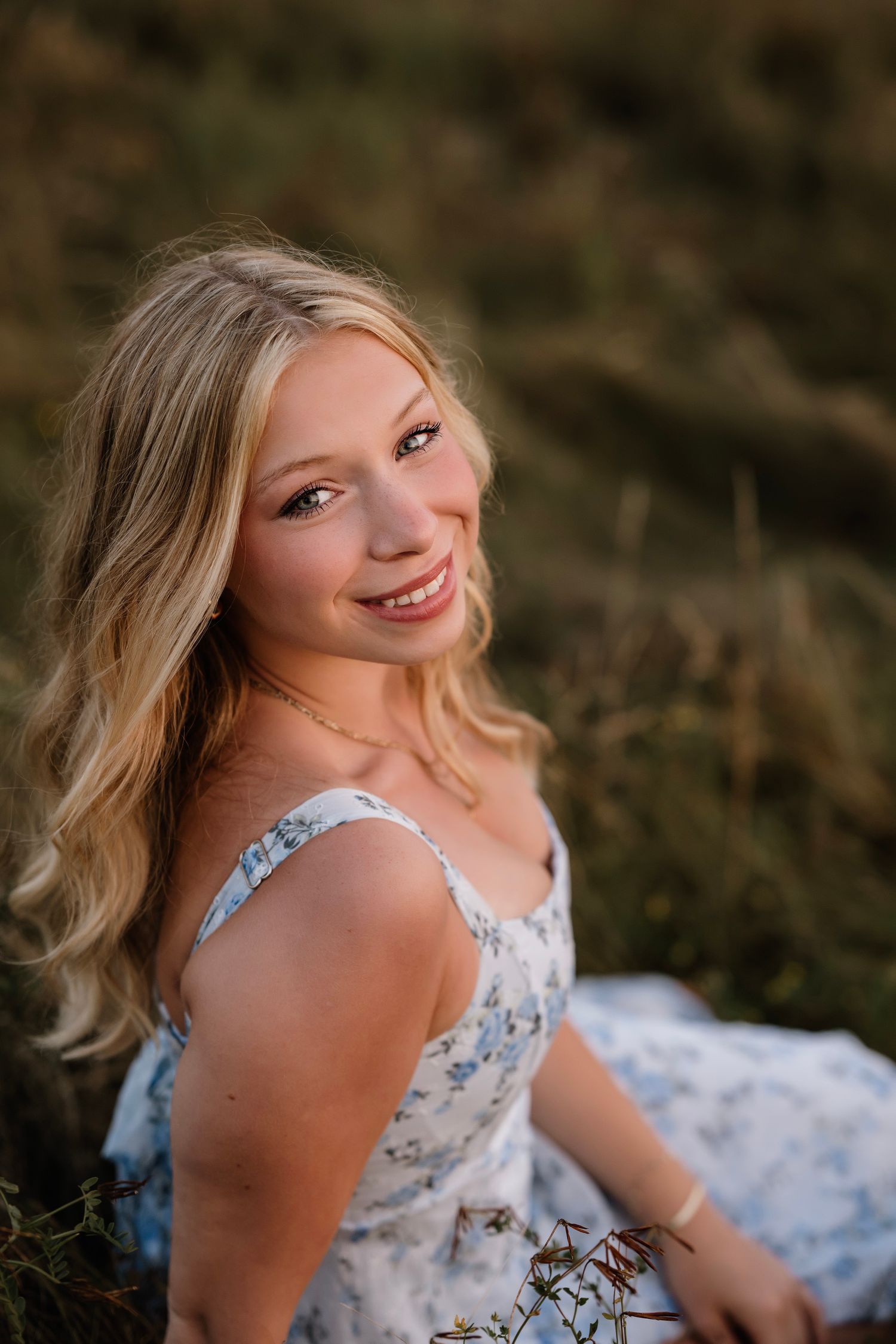 Smiling young woman with long wavy blonde hair in a blue floral sundress sits in a grassy field, glowing in warm golden-hour light.