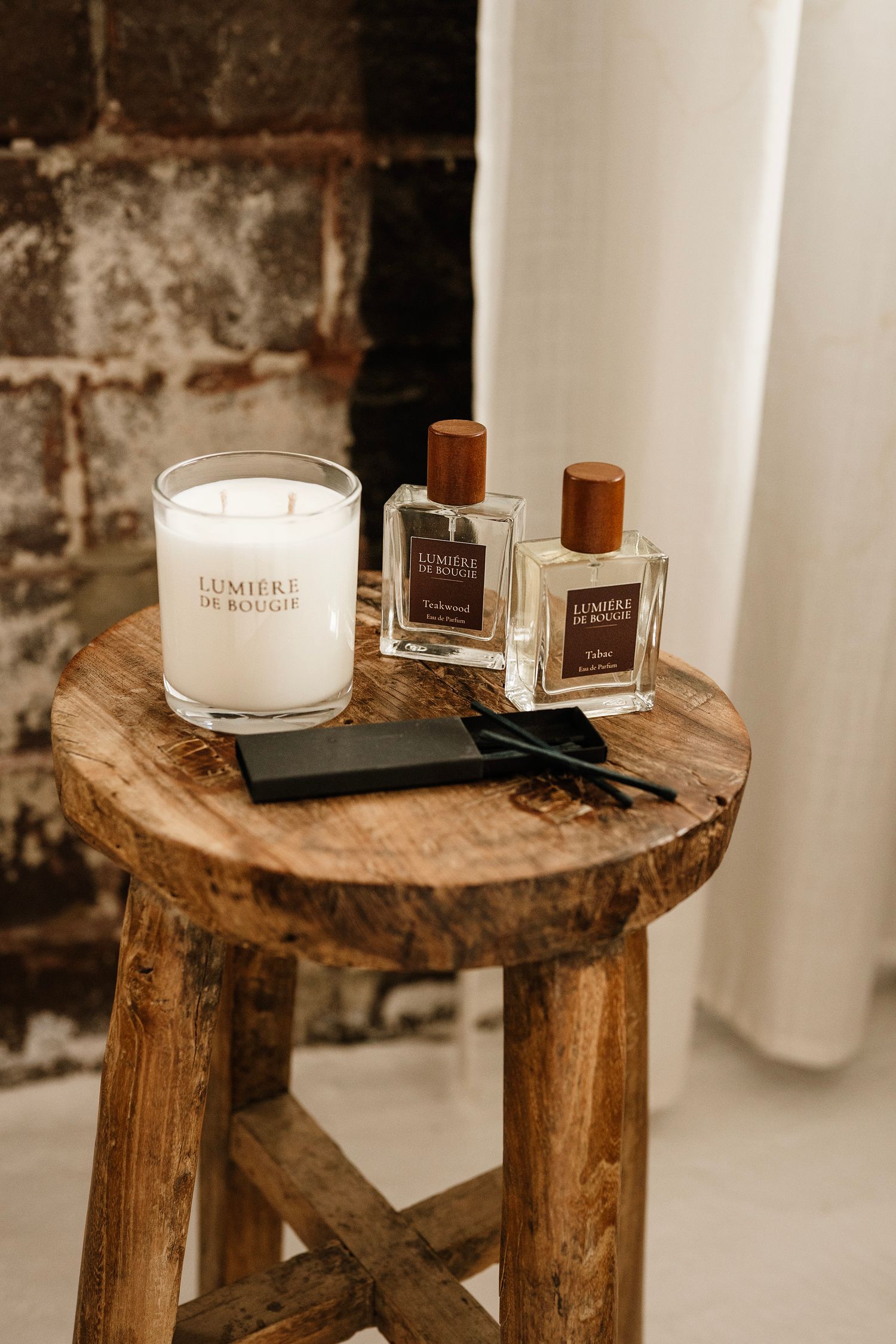 Lumiere De Bougie candle and two perfume bottles displayed on a rustic wooden stool against a brick wall.