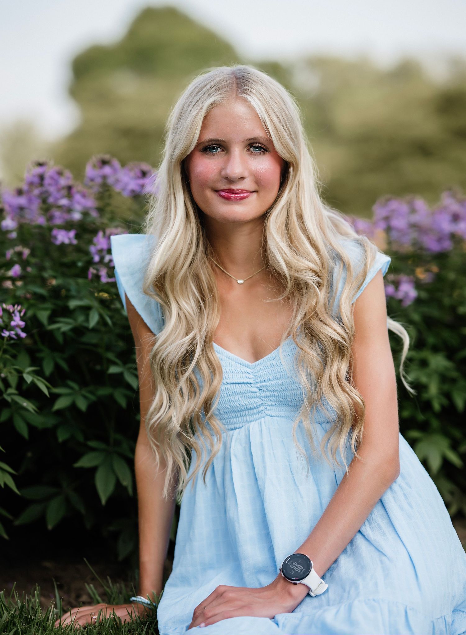 A person in a light blue dress poses outdoors against purple wildflowers on a summer day.