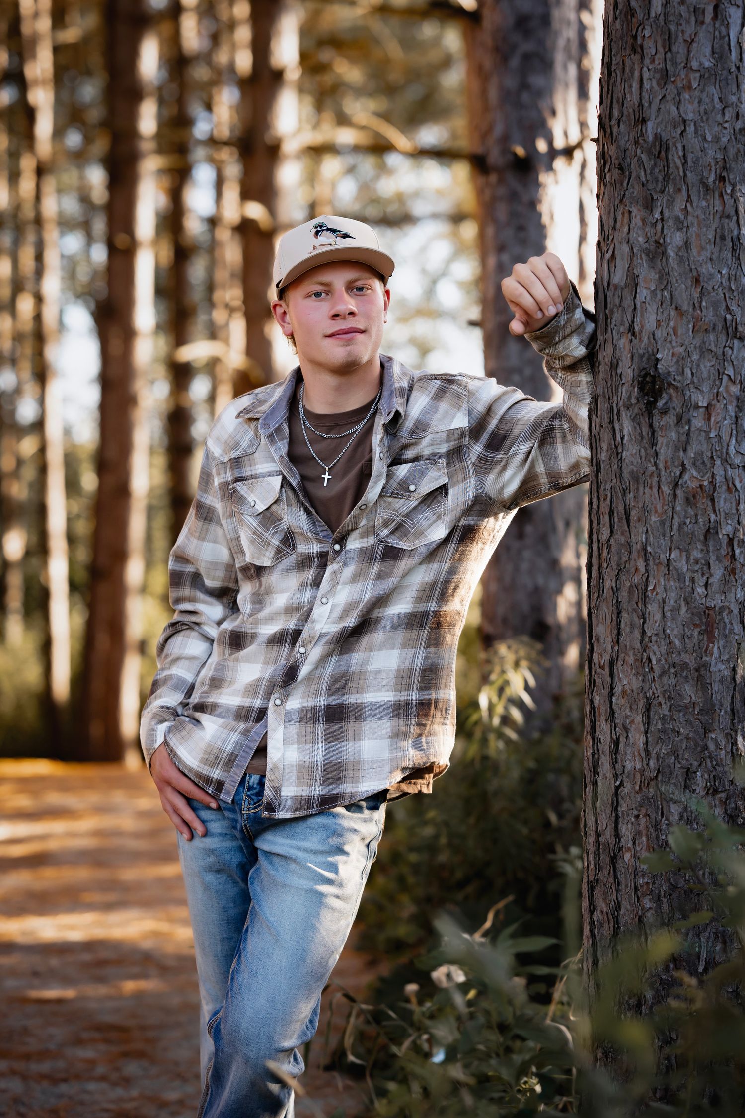 Person in plaid flannel shirt and baseball cap leans casually against tree trunk in sunlit forest setting.