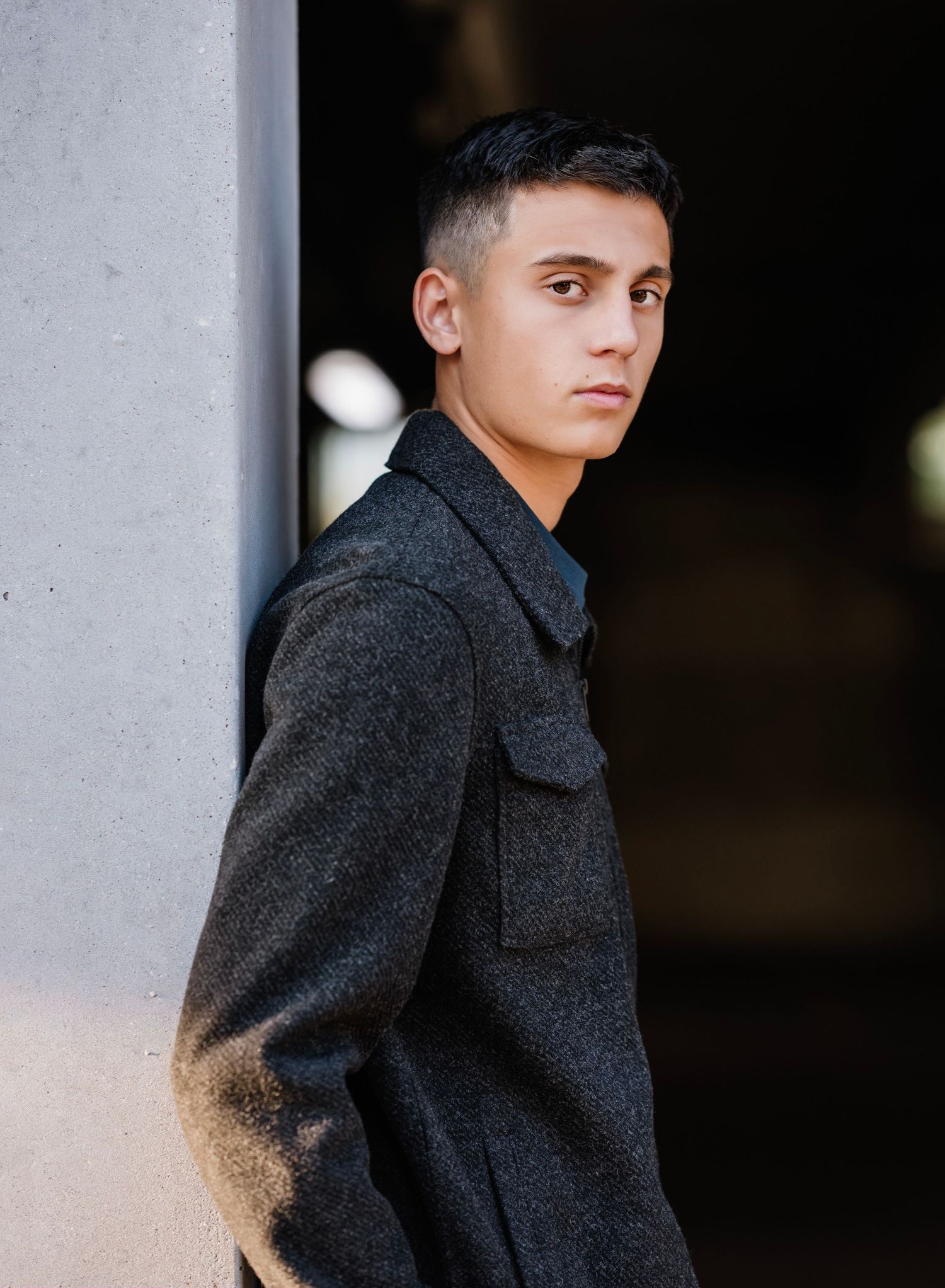Young person in a dark wool coat poses against a concrete wall in dramatic lighting with a contemplative expression.