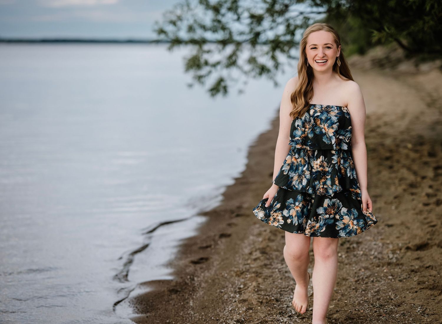 A person in a floral dress walks along a sandy beach shoreline with water and trees in the background.