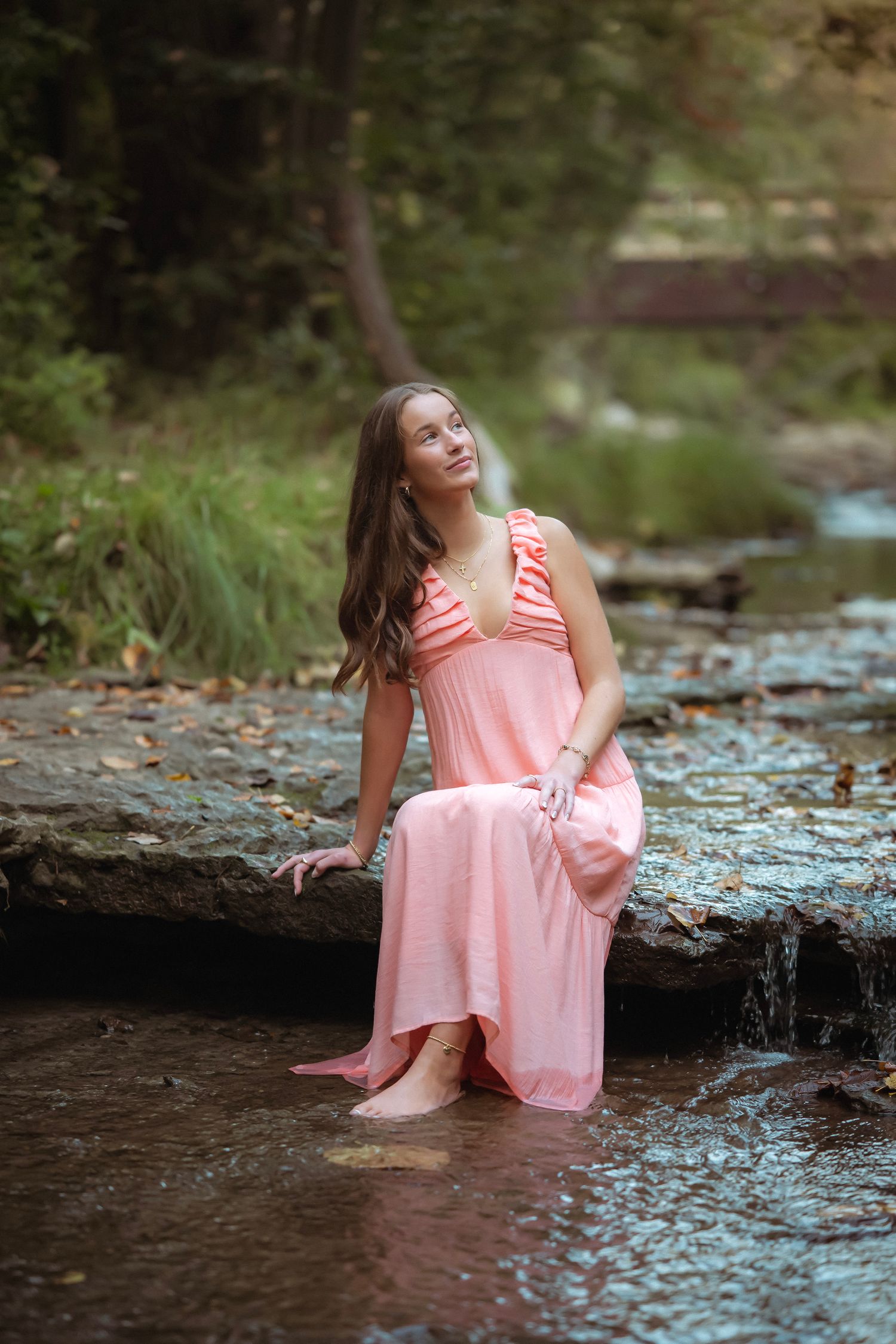 A person in a flowing pink dress sits peacefully on rocks near a forest stream on a summer day.