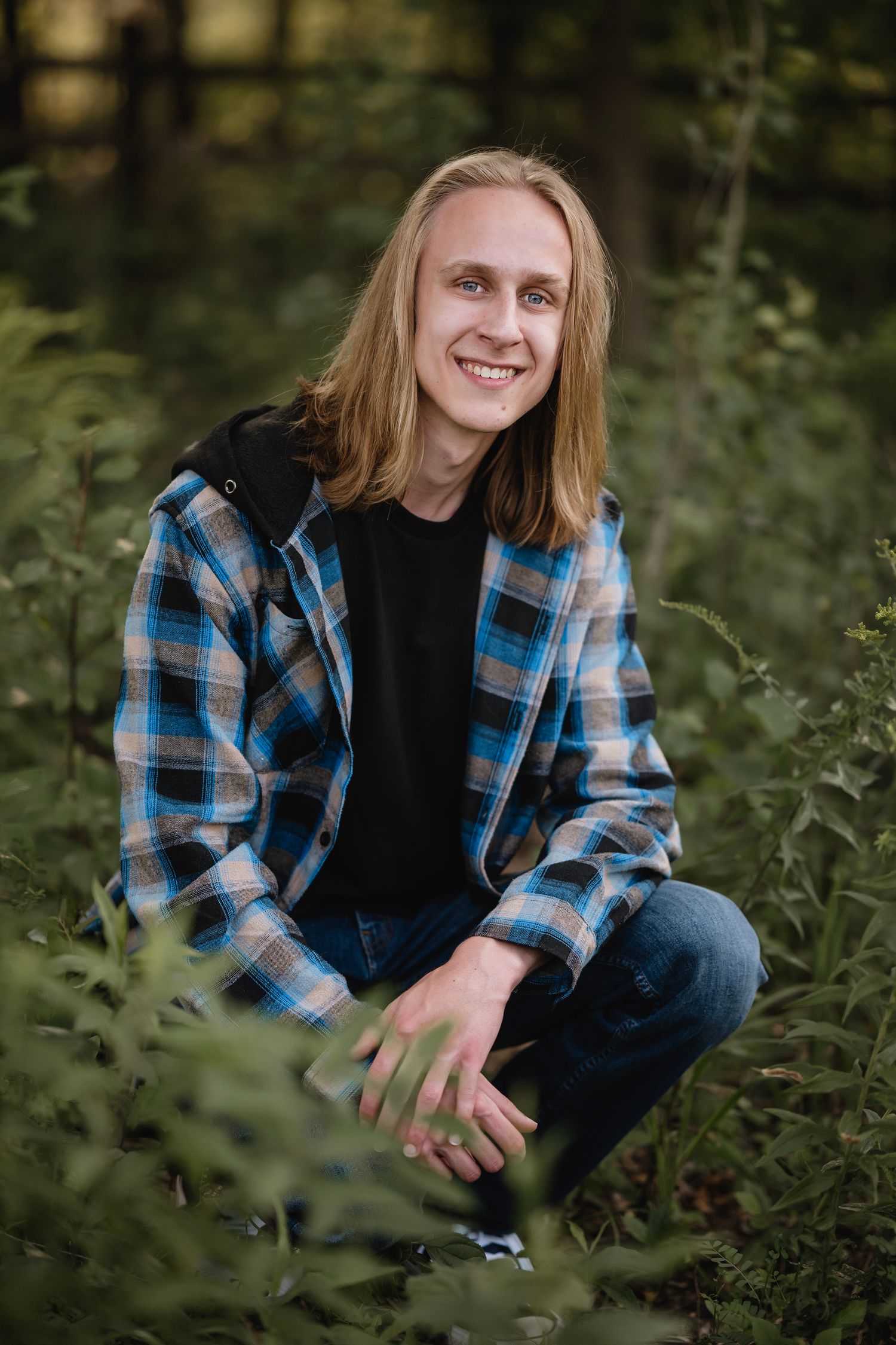 Young man with long blonde hair smiling in forest, wearing plaid shirt and jeans, crouching among green plants.