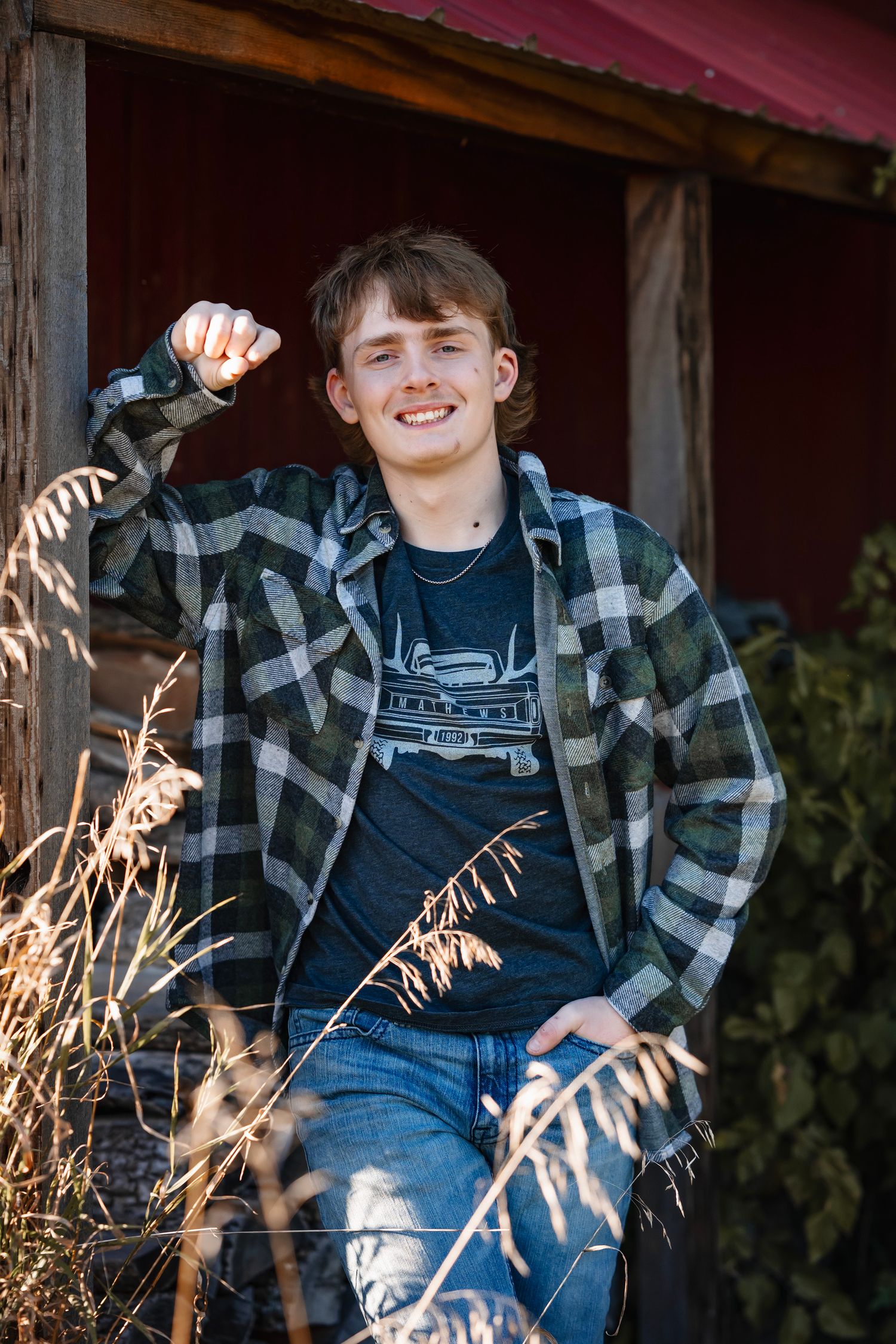 Child in blue denim overalls and plaid shirt sits on rustic wooden porch with tall grass in foreground.