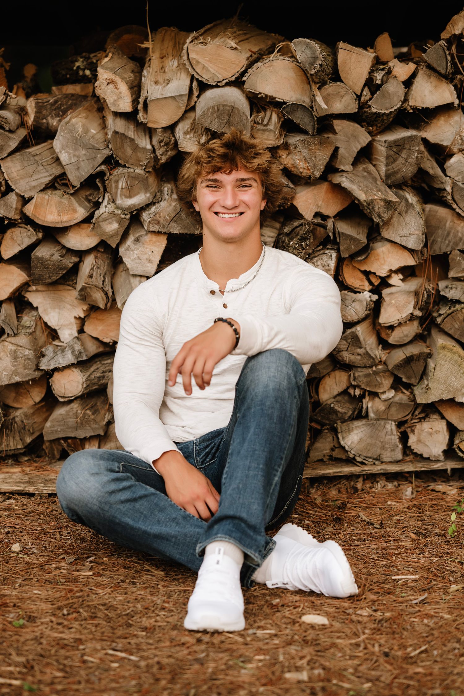 Smiling young man in white henley and jeans sitting cross-legged in front of a stacked firewood pile outdoors.