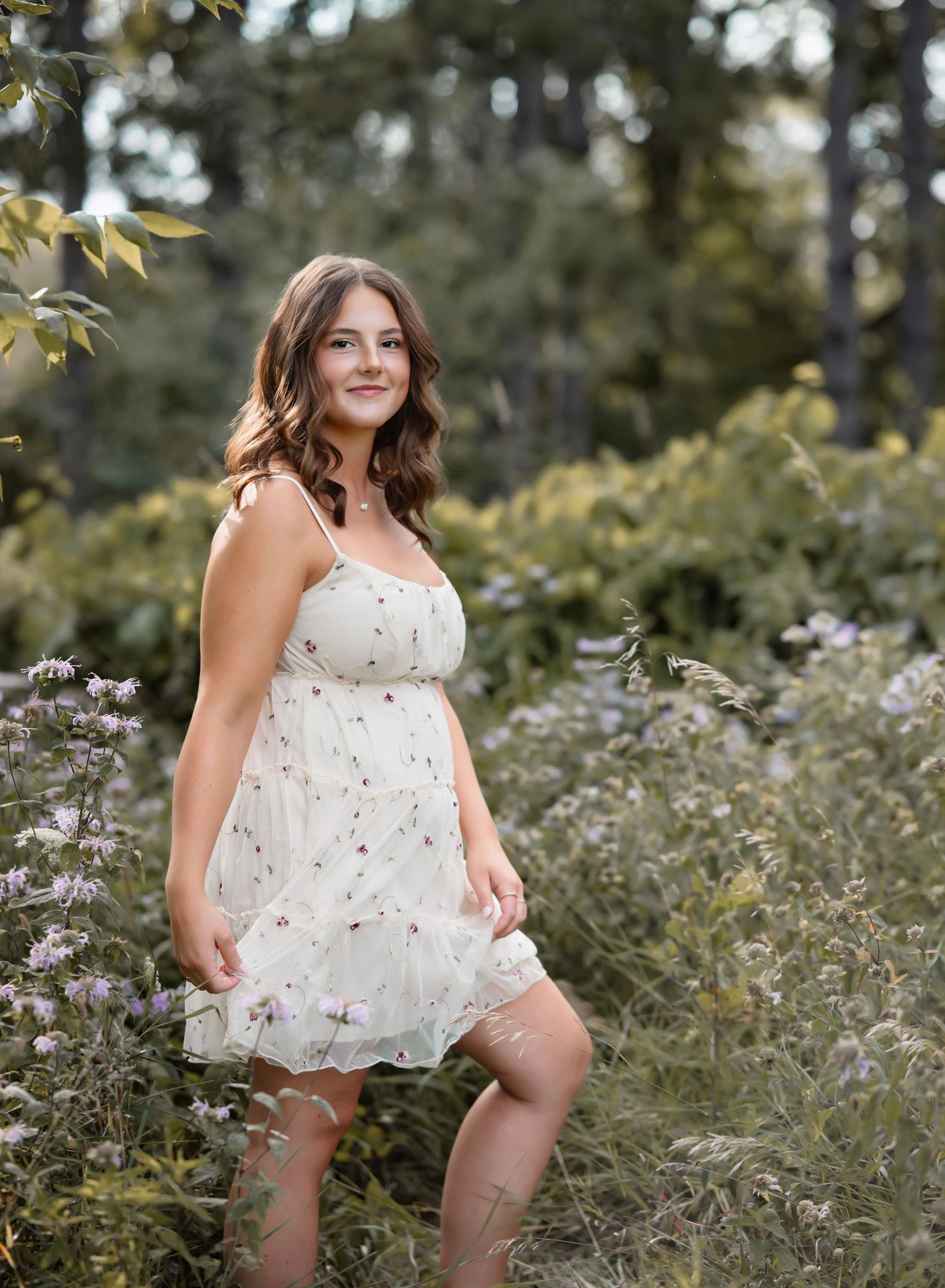 A person in a white floral sundress poses outdoors among wildflowers and greenery on a sunny summer day.